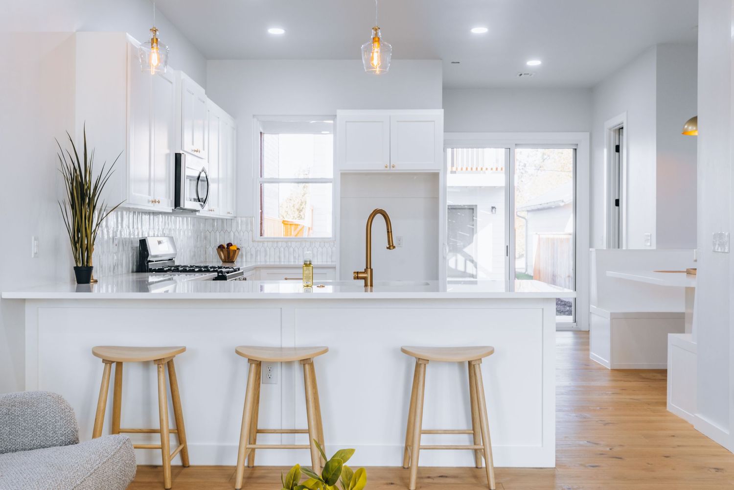 Modern white kitchen with wooden bar stools, gold pendant lights, and a bronze faucet features bright natural lighting.