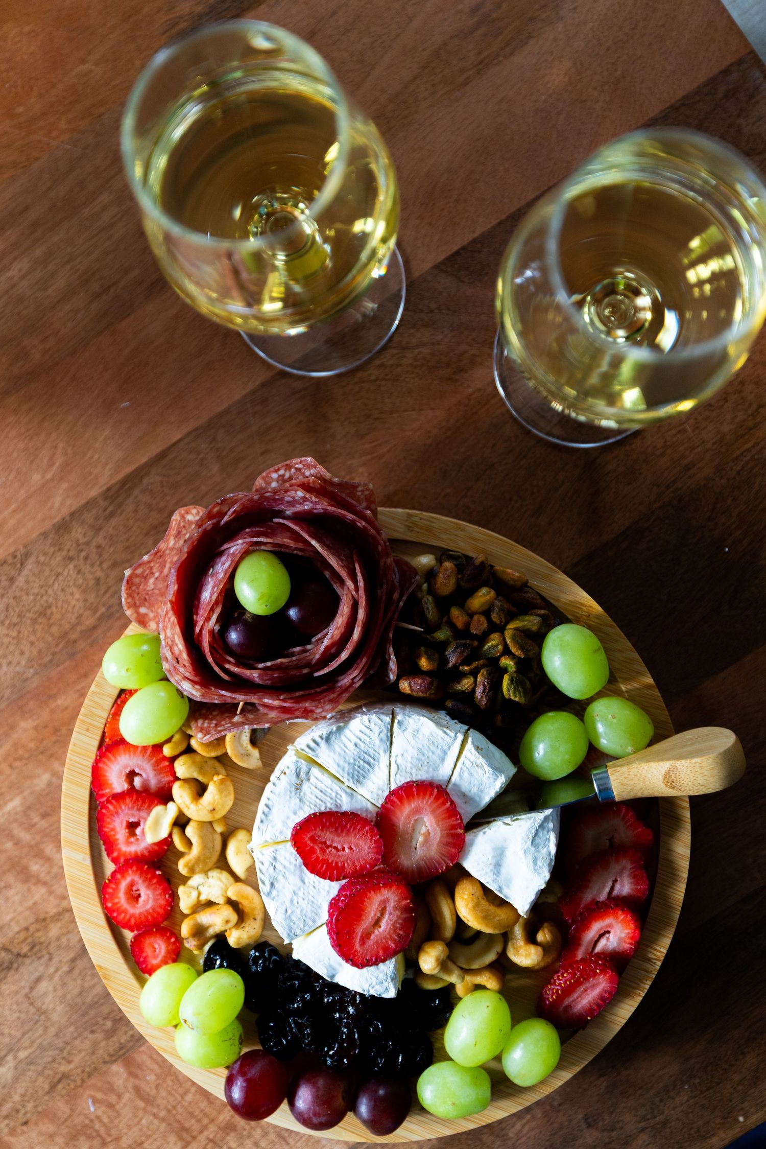 Overhead view of charcuterie board with wine, brie cheese, grapes, strawberries, cashews and prosciutto on wooden table.