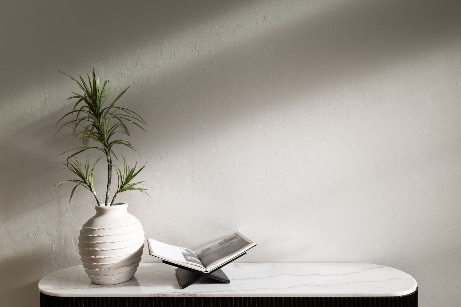 Minimalist white vase with plant and book on light surface against neutral wall with dramatic lighting.