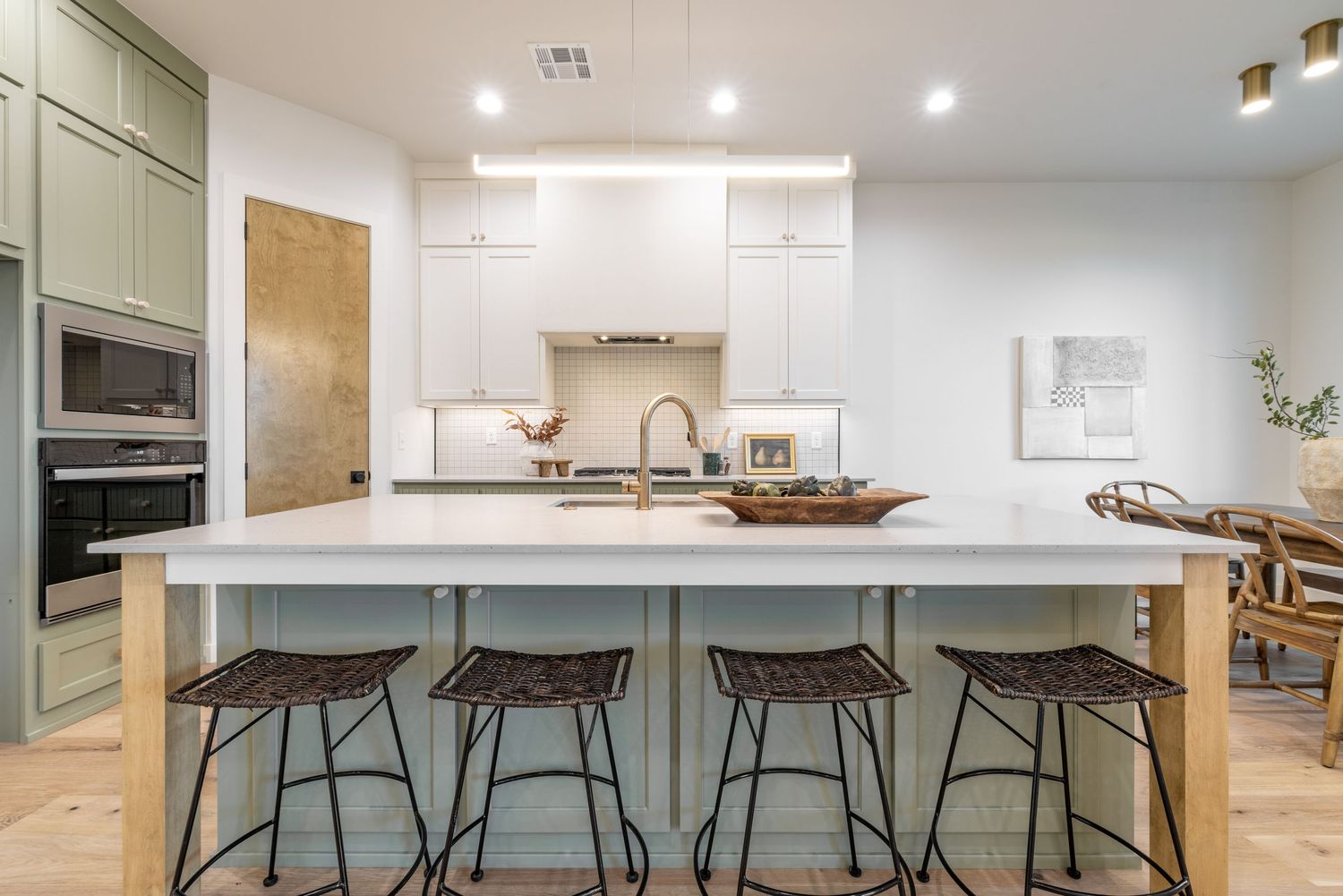 Modern kitchen features sage green cabinets, white quartz island with metal barstools, and warm hardwood floors.