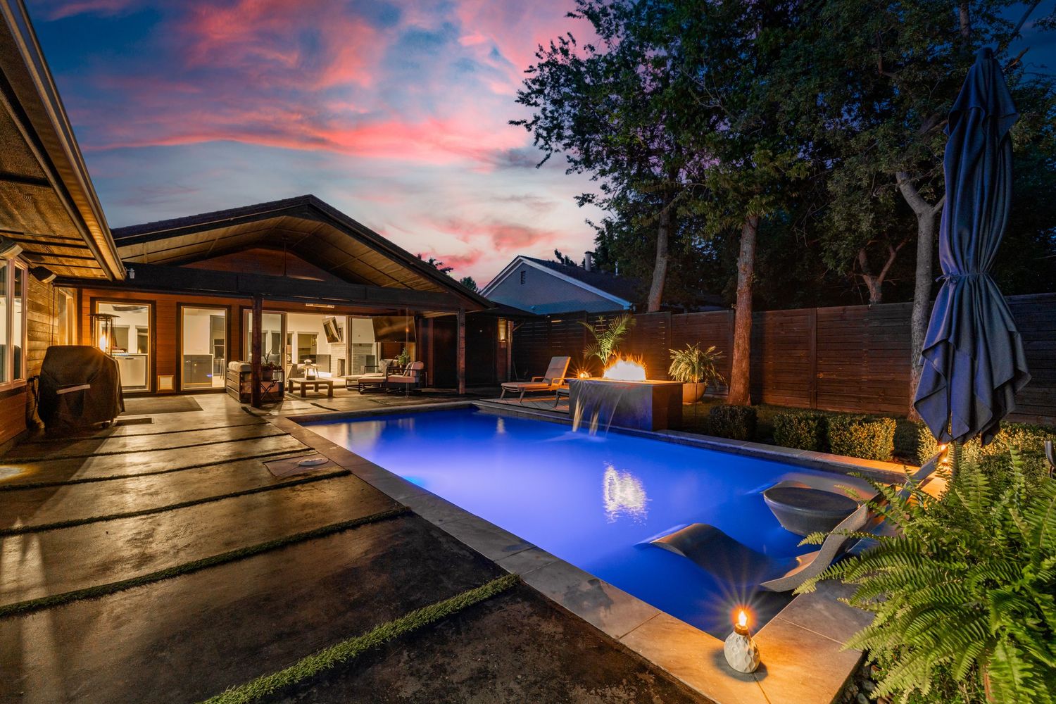 Luxurious backyard with illuminated blue pool and modern patio under vibrant sunset sky.