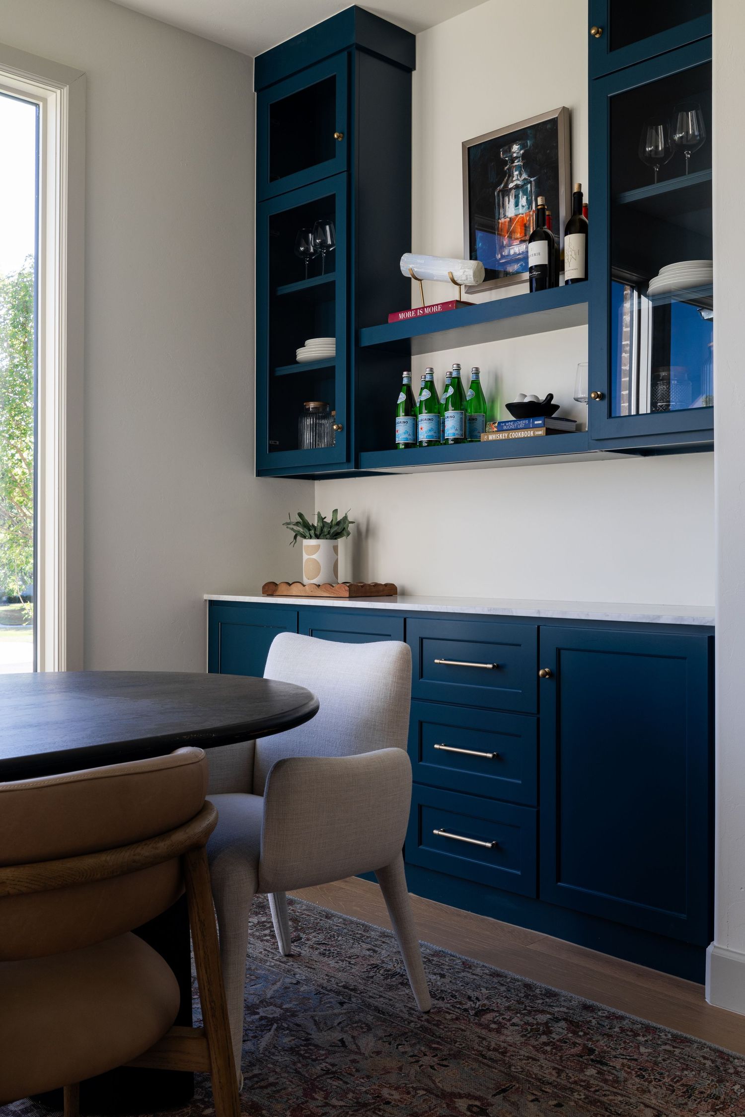 Modern dining area with navy blue built-in cabinets, round table, upholstered chairs and display shelves for bottles.