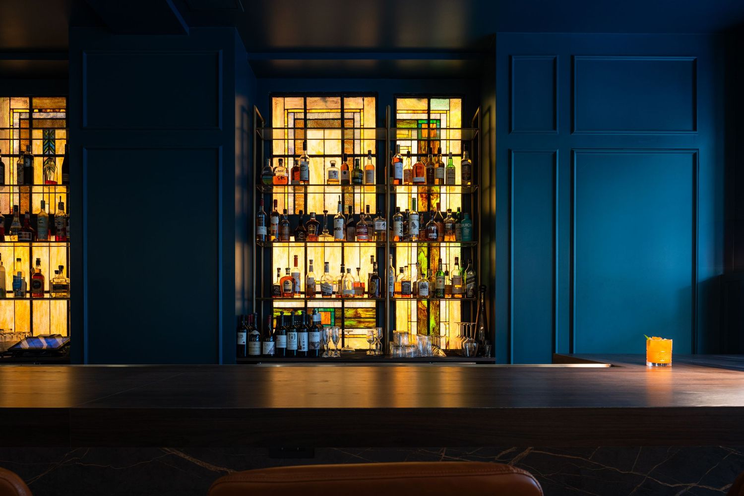 Illuminated liquor bottles glow warmly through windows at night, creating an inviting bar display against dark blue walls.