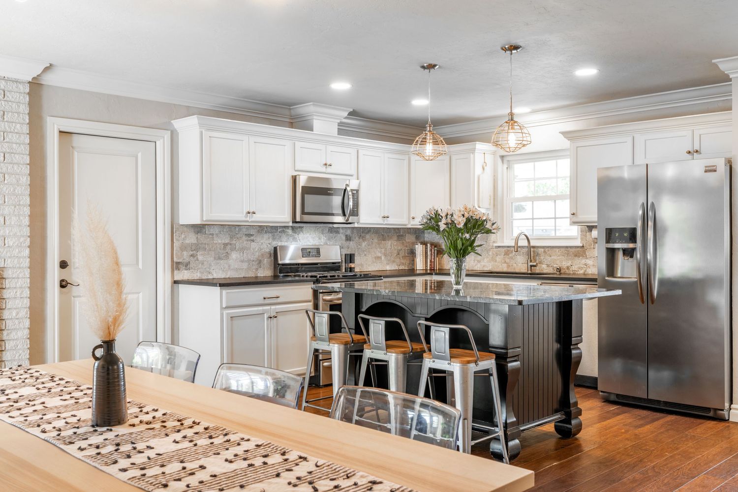 Modern white kitchen with stainless steel appliances, black island, gray barstools and pendant lighting over counter.