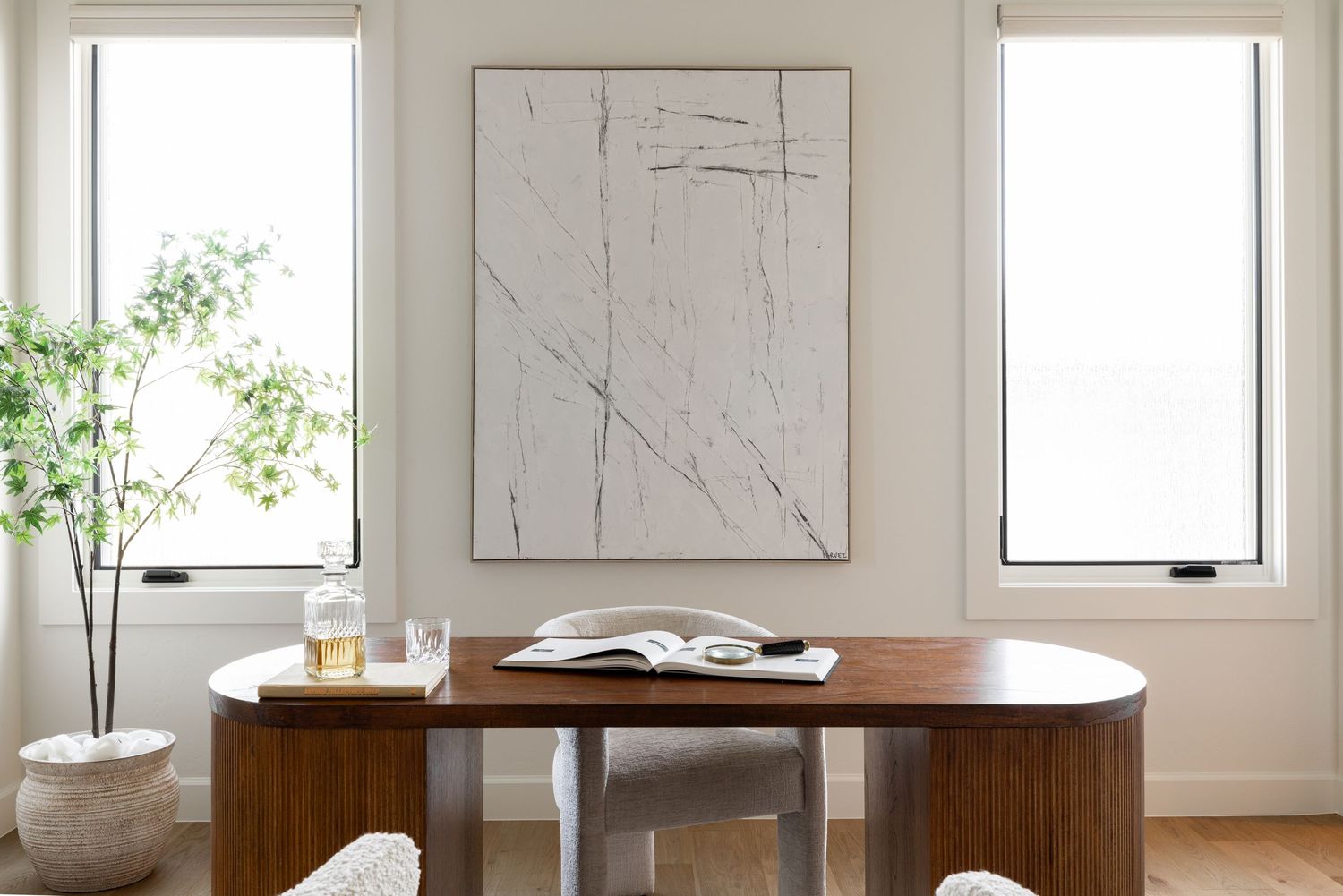 Minimalist dining area with oval wooden table, clear chairs, abstract white artwork and tall windows.
