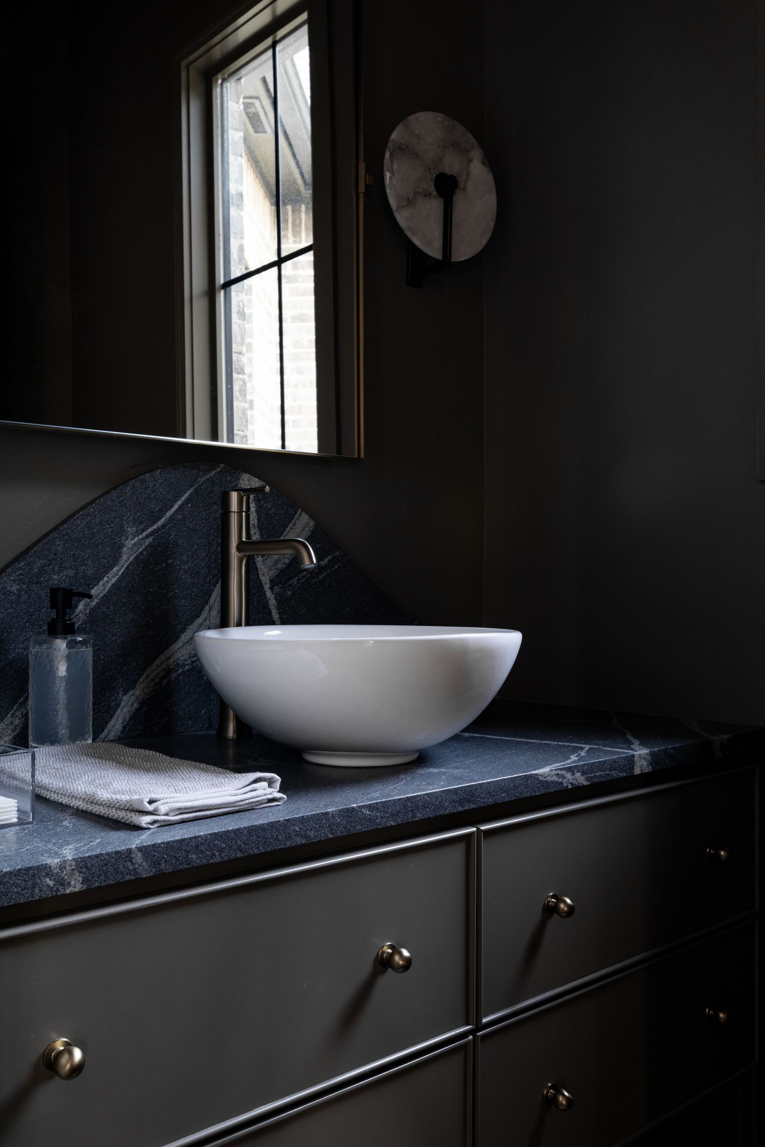 Modern bathroom vanity features white vessel sink on black marble countertop with dark cabinets and tall window.
