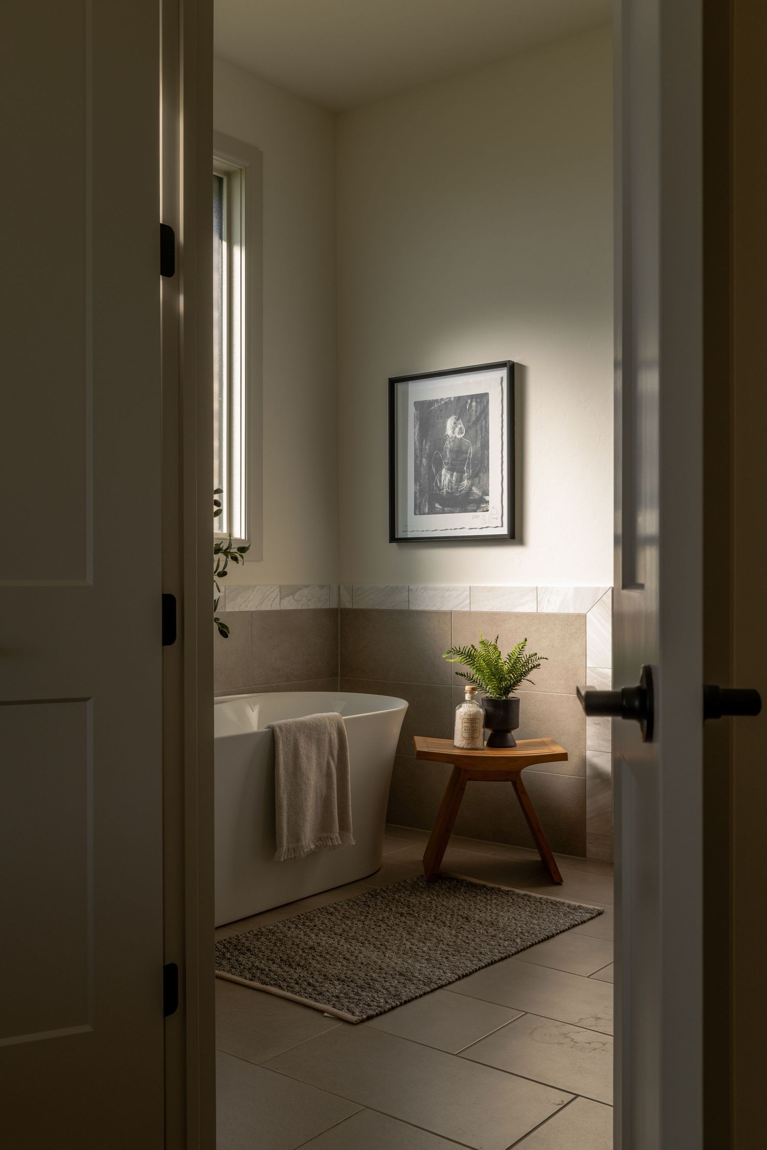 Minimalist bathroom interior with freestanding tub, wooden stool, houseplant and framed artwork viewed through doorway.