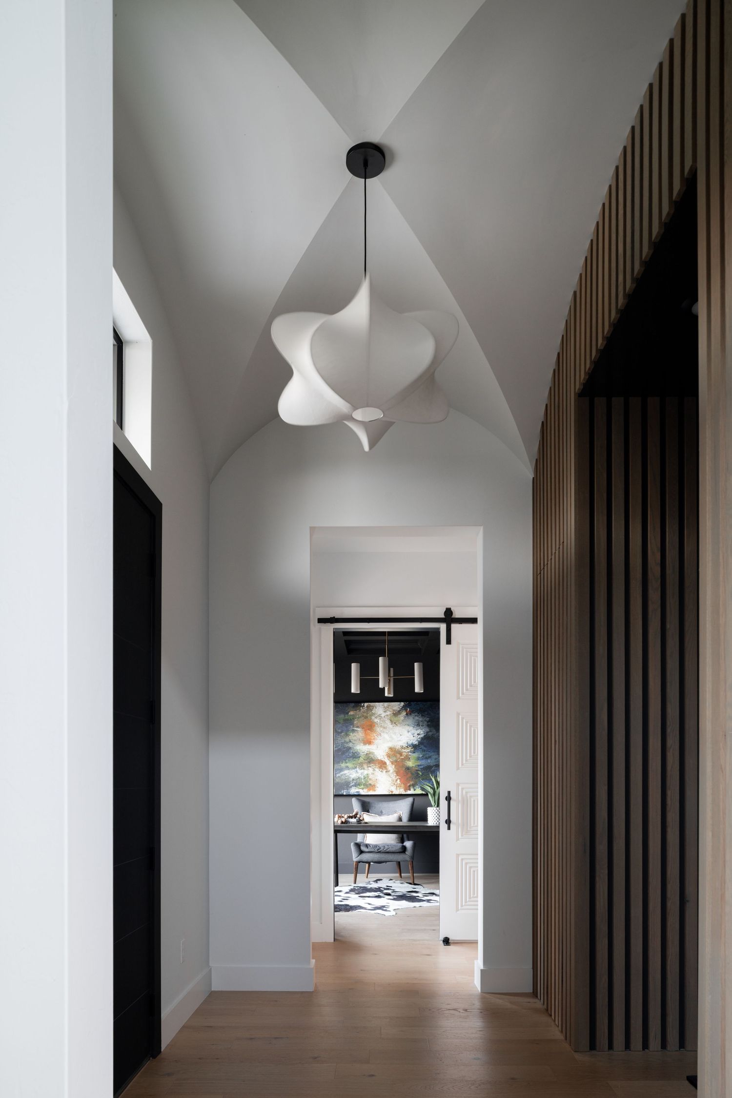 Modern hallway with sculptural white pendant light, wooden slat wall detail, and glimpse into room with contemporary artwork.