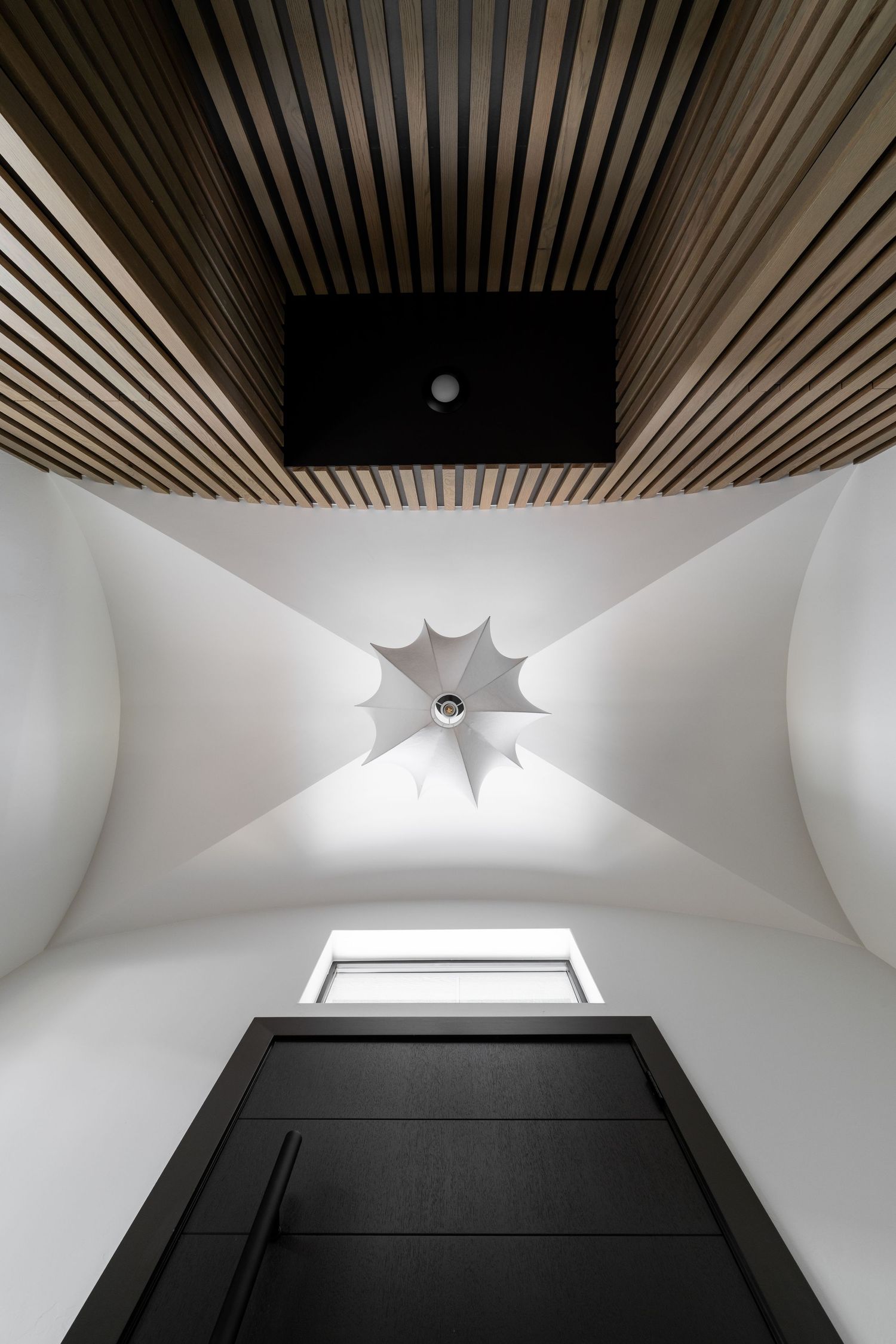 Looking up at a modern ceiling with wood slats, geometric white curves, and a star-shaped light fixture above a black door.