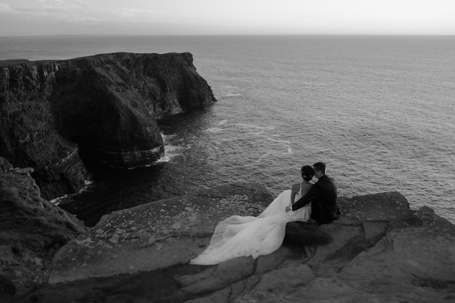 A black and white wedding photo captured on dramatic coastal cliffs overlooking the ocean.