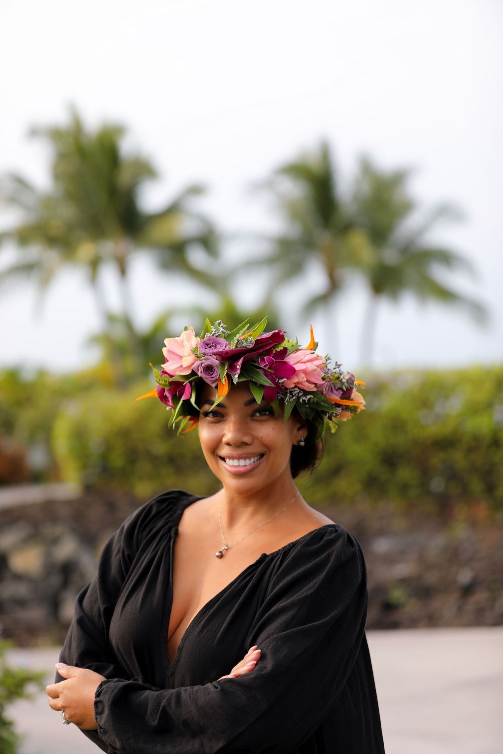 Person wearing a vibrant tropical flower crown and black dress smiles outdoors with palm trees in the background.