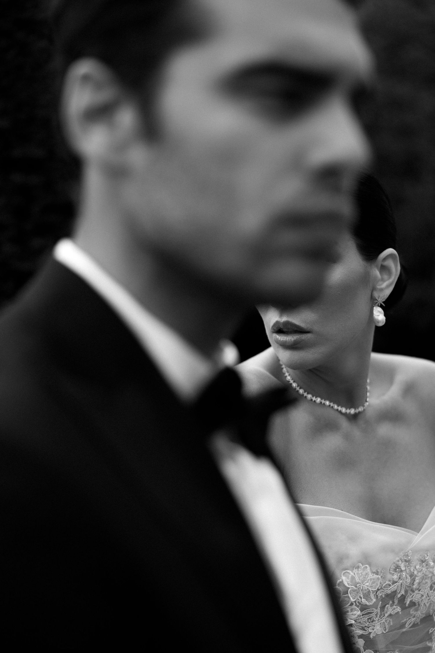 Artistic black and white wedding portrait showing the bride in focus with pearl necklace behind the groom's silhouette.