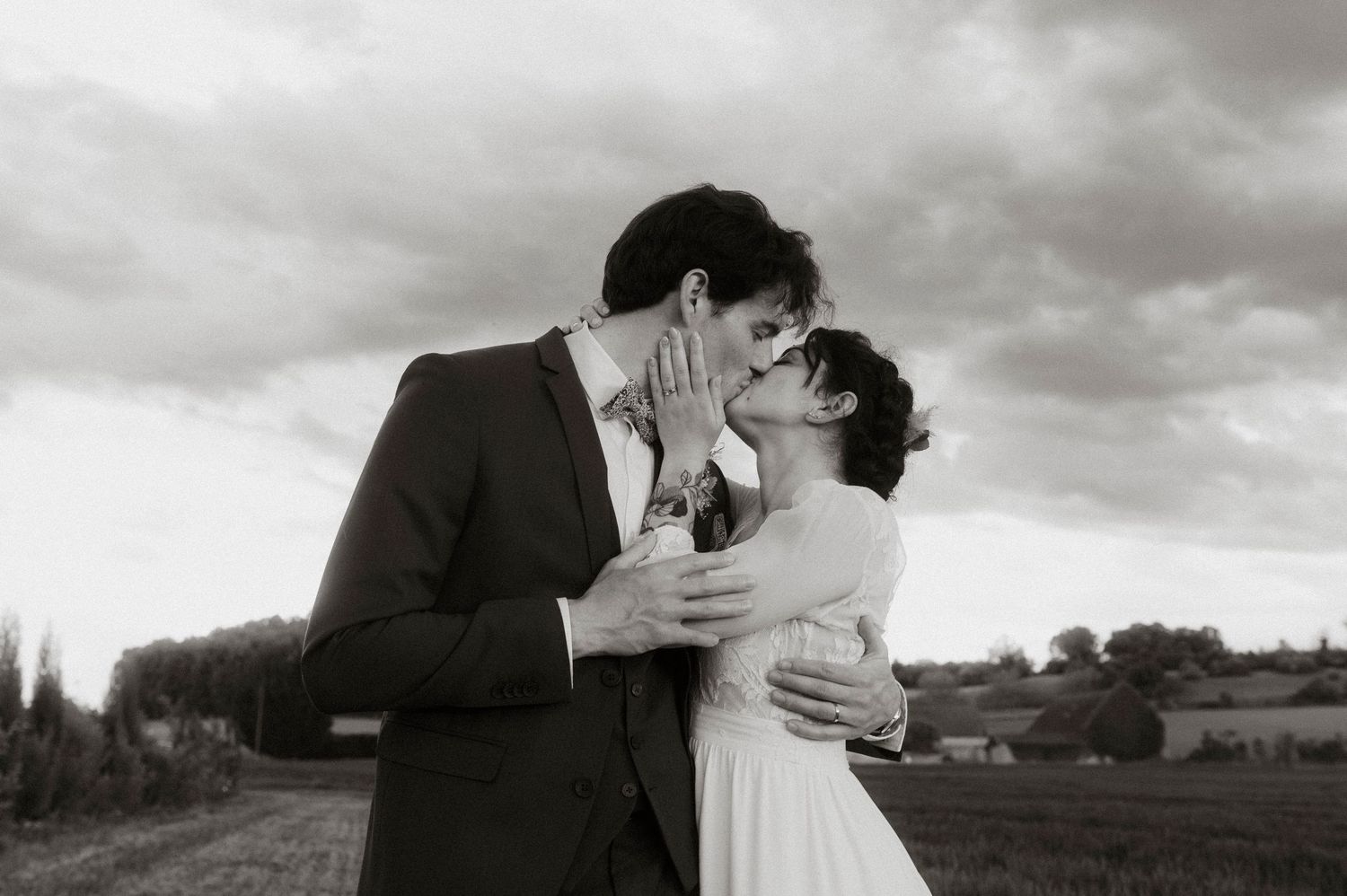 A couple in vintage formal attire share a romantic embrace and kiss in a countryside setting under dramatic cloudy skies.