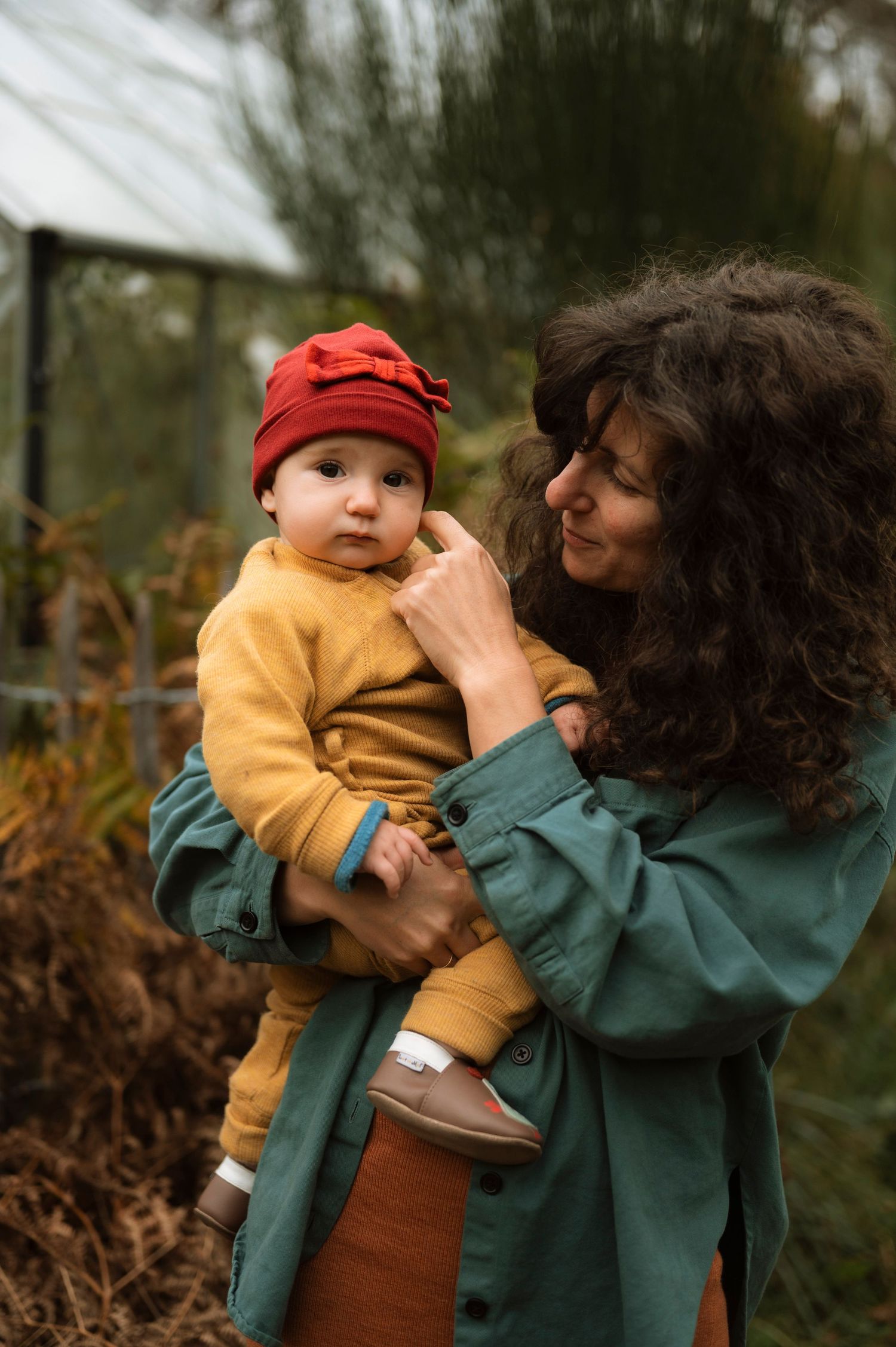 A person in a green jacket holds a small baby wearing a yellow sweater and red hat outdoors on an autumn day.