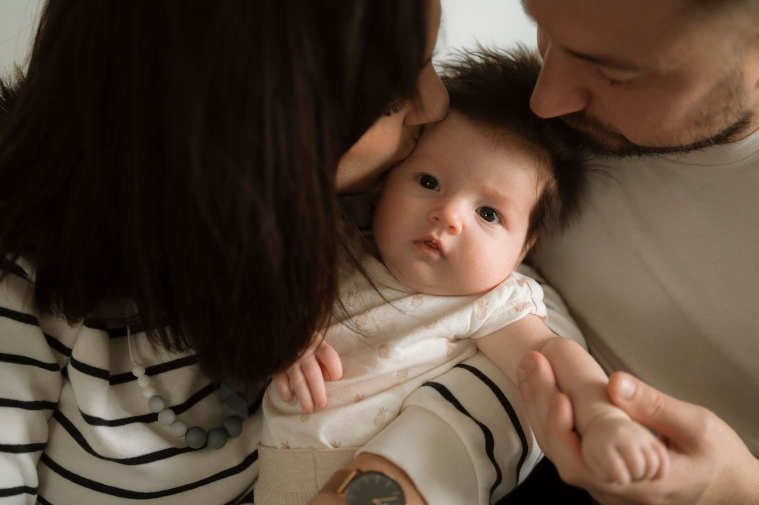 A baby in a white onesie lies between two people in a tender family moment.