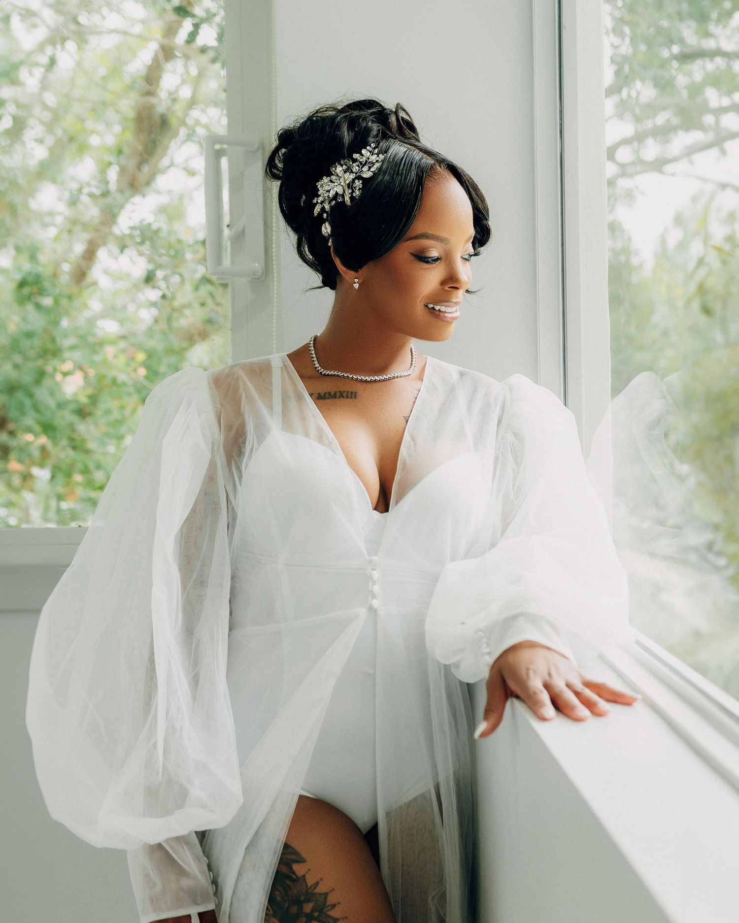 A bride in a white dress with billowing sleeves poses elegantly by a window.