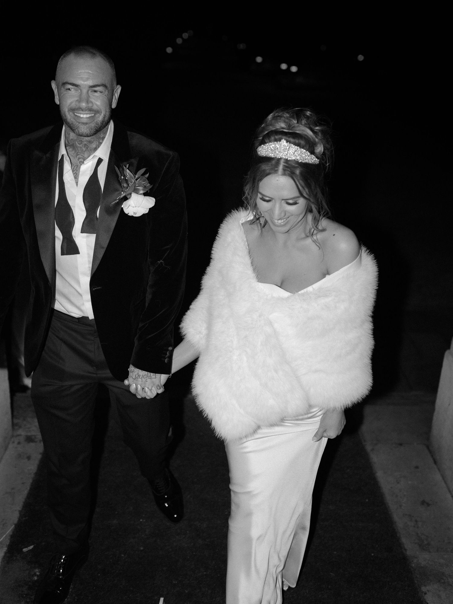 A couple holds hands at a formal event, with the groom in a tux and bride wearing a white gown and fur wrap.