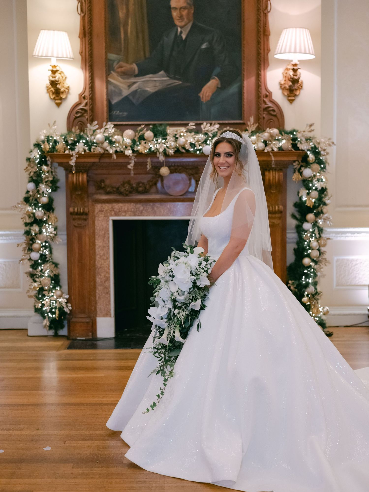 Beautiful bride in flowing white ballgown poses by ornate fireplace decorated with white floral arch and portrait above mantel.