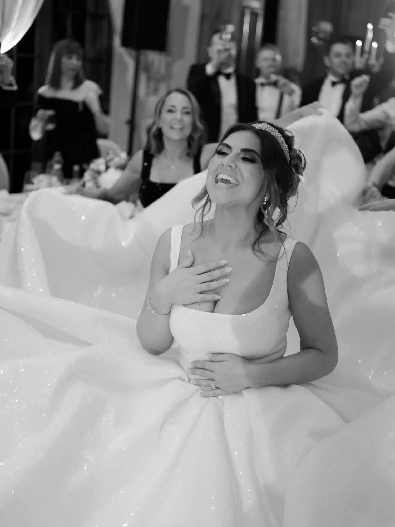 A bride in a voluminous white ball gown laughs joyfully during a wedding celebration while seated.