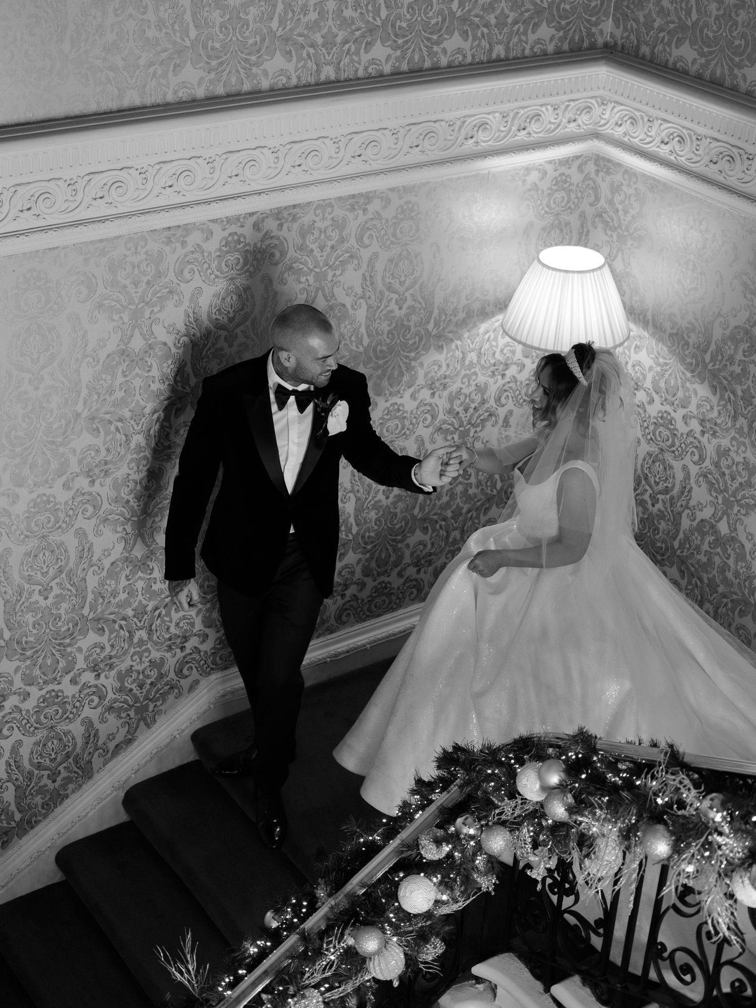 A black and white wedding photograph on elegant staircase with ornate wallpaper and glowing lamp in a classic setting.