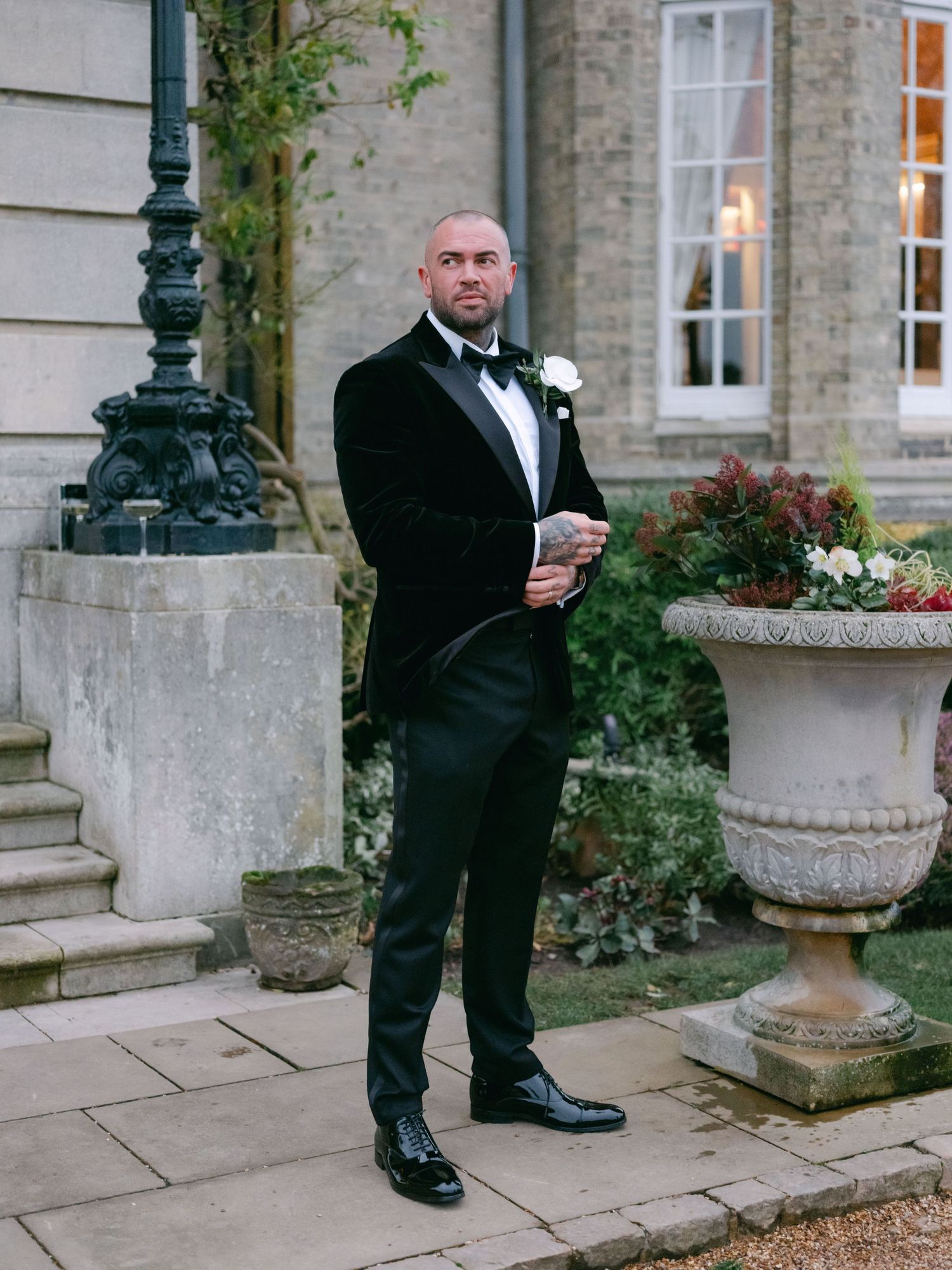 A well-dressed person in a black tuxedo holds a drink while standing beside elegant stone planters at a formal event.