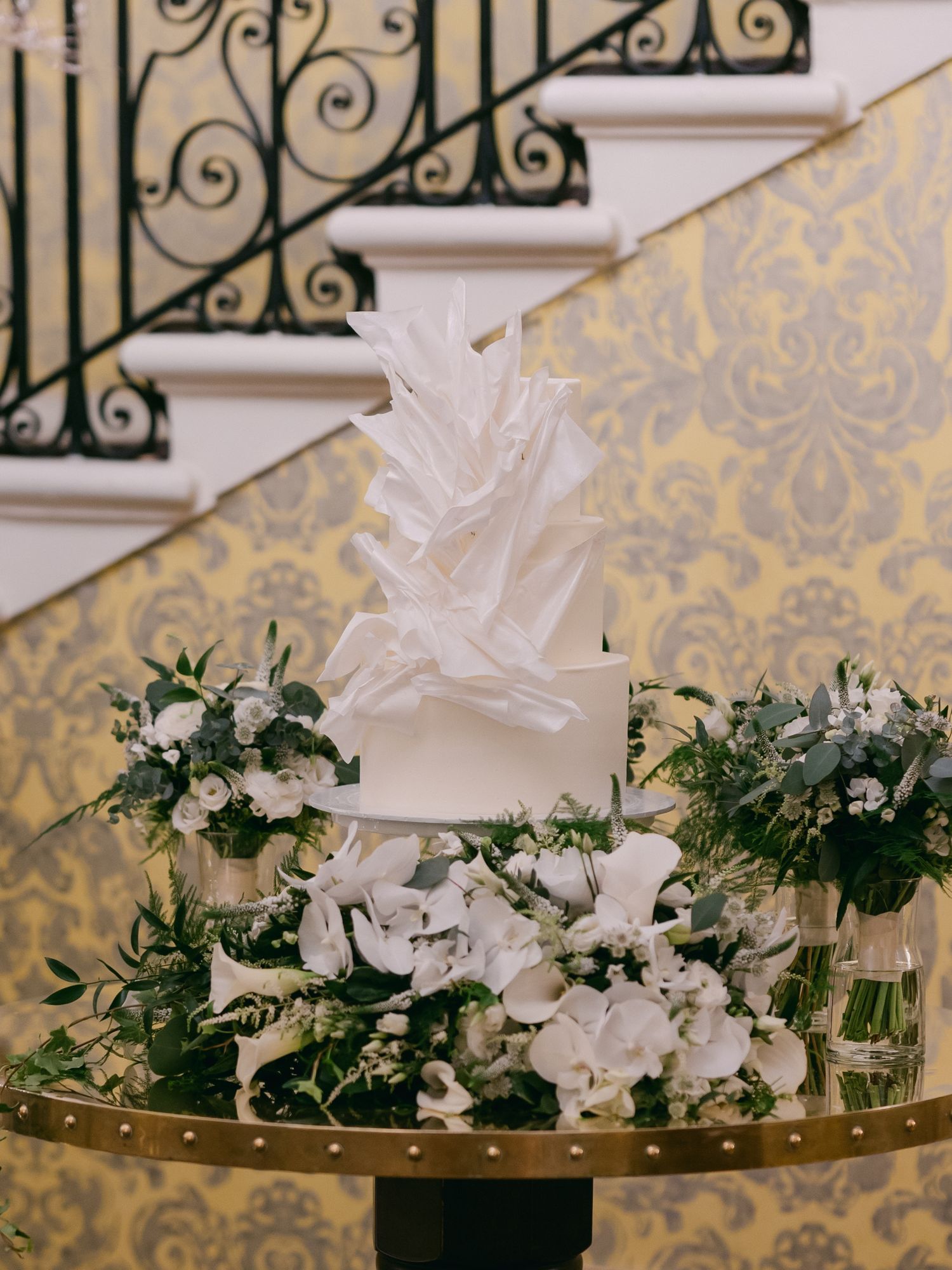 White floral arrangements with greenery sit atop a glass table with damask wallpaper in the background.