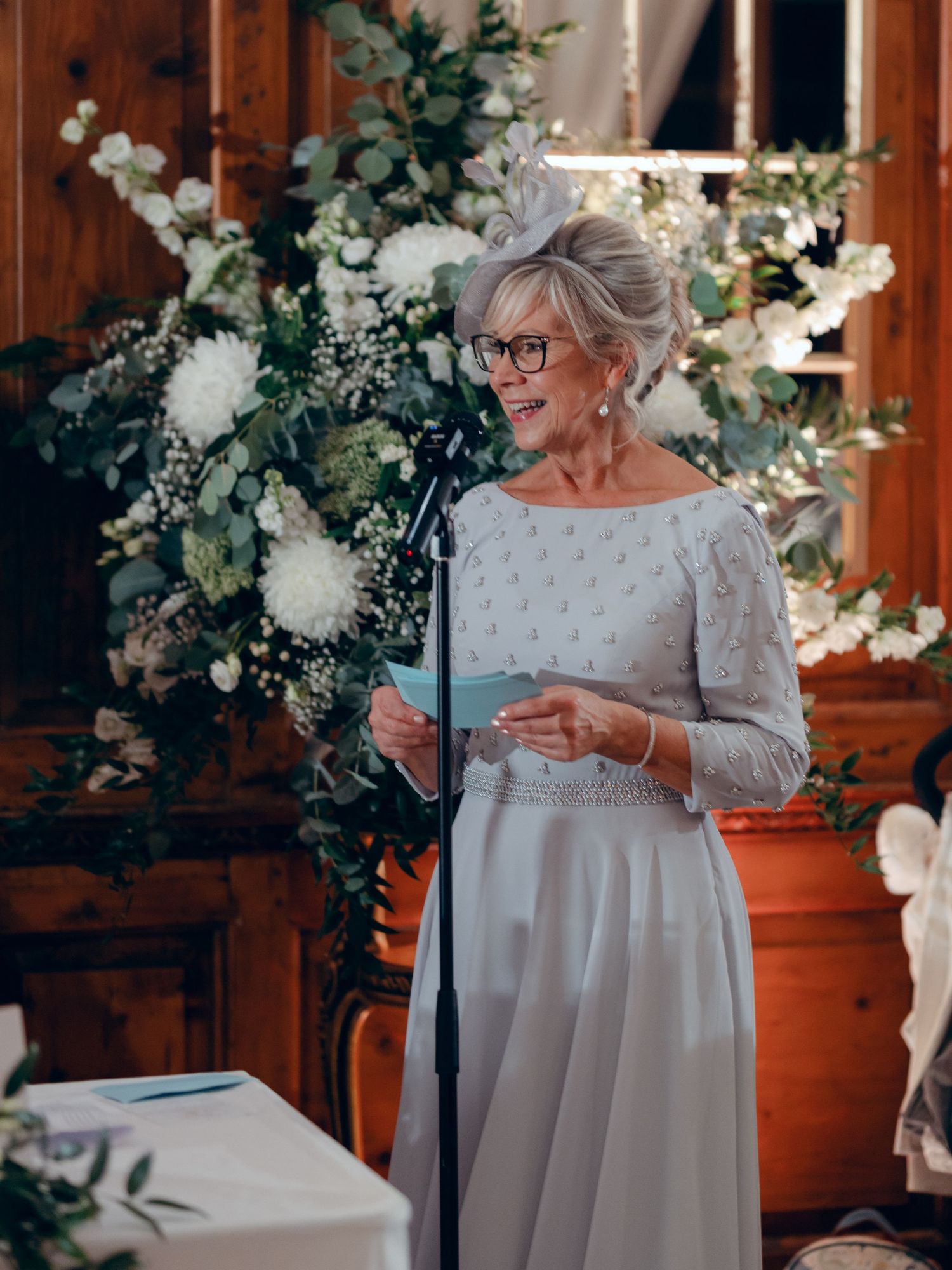 Speaker wearing a light gray lace dress stands at podium with elegant white floral arrangements and greenery in background.