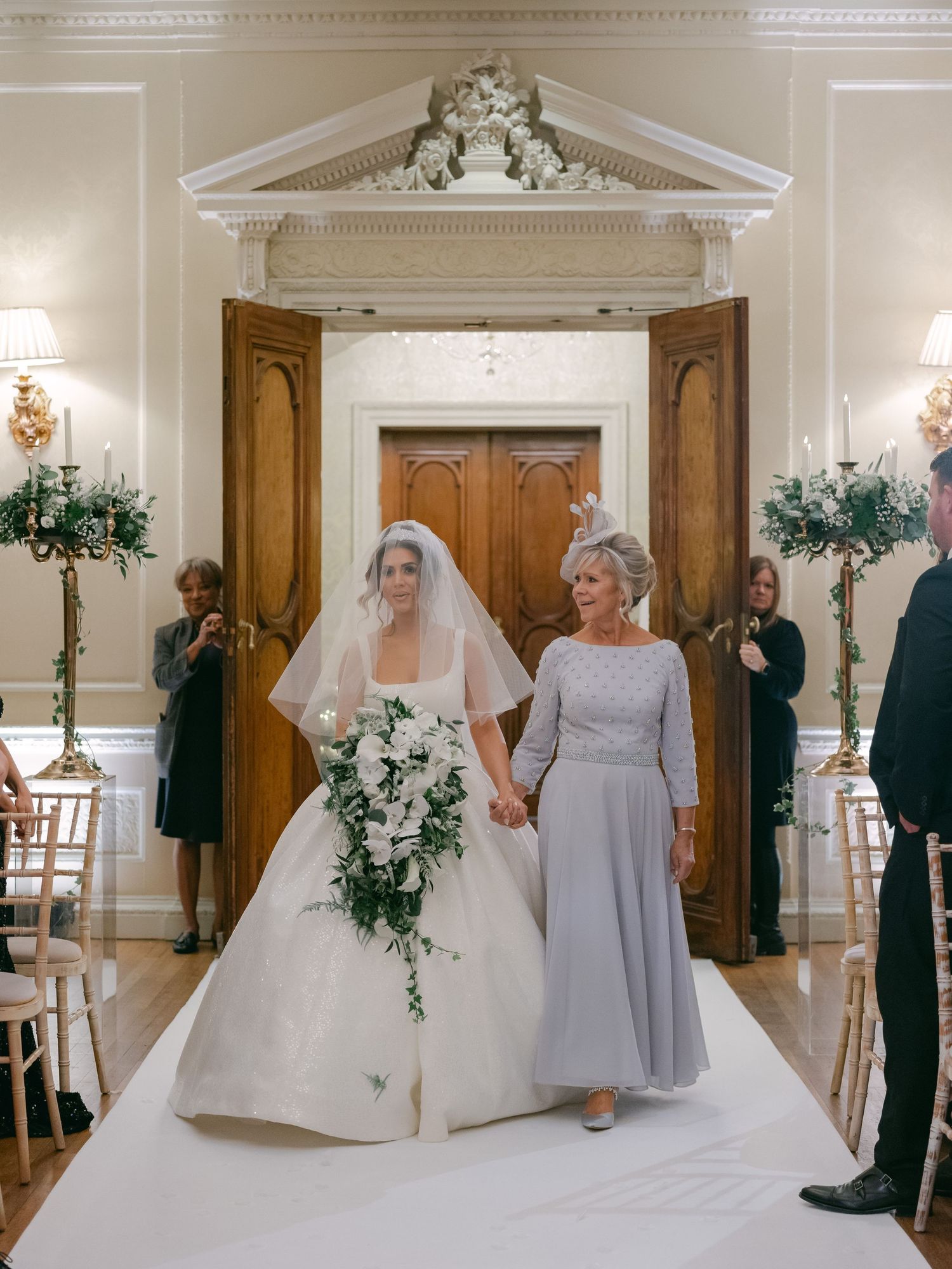 Two wedding attendees walk down an elegant white aisle with grand wooden doors and floral arrangements in the background.