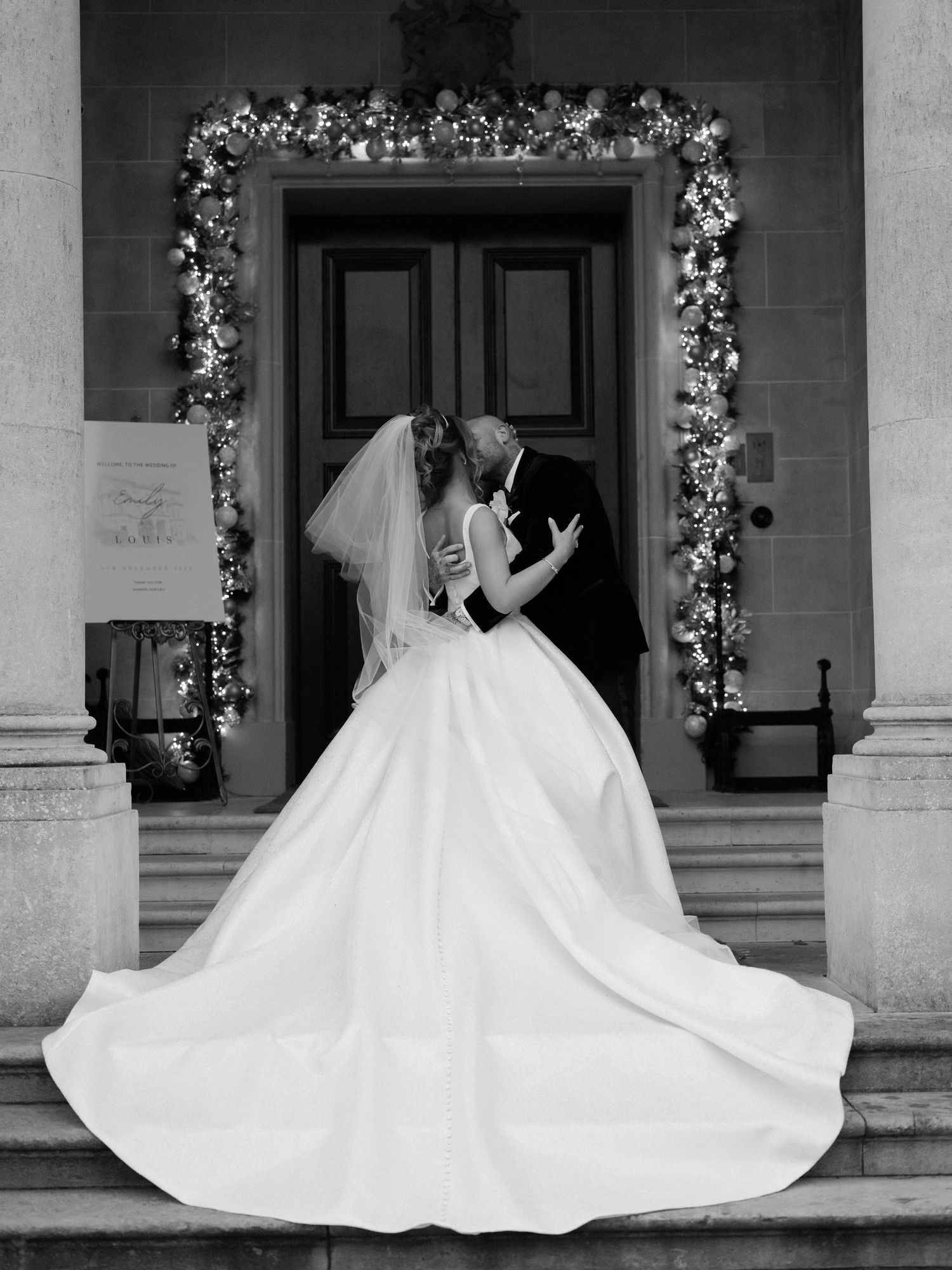 A romantic black and white wedding photo showing a couple kissing in a doorway adorned with floral decorations.