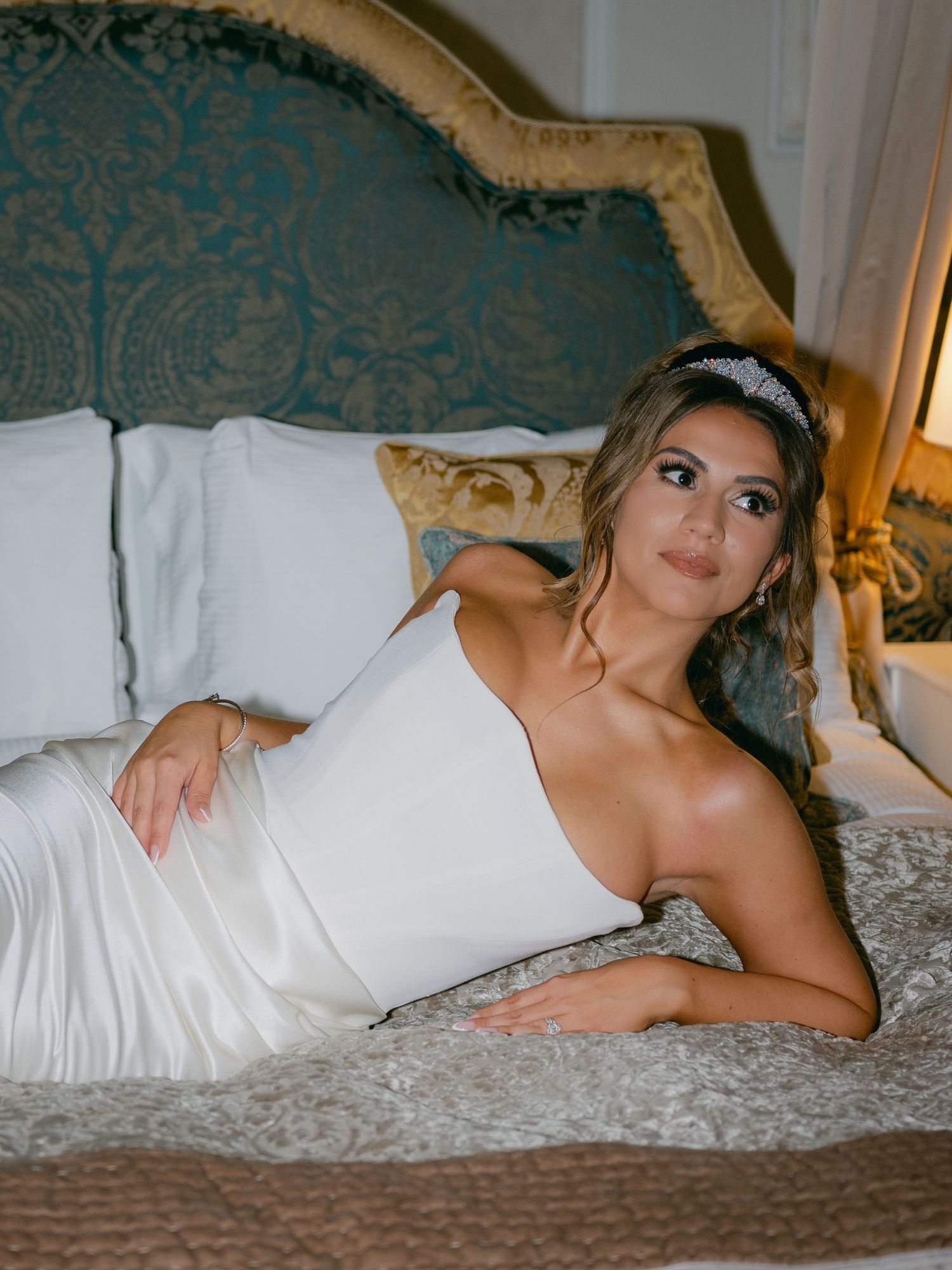 A figure in a white dress reclines on a patterned bed with ornate teal headboard in boudoir photography series.