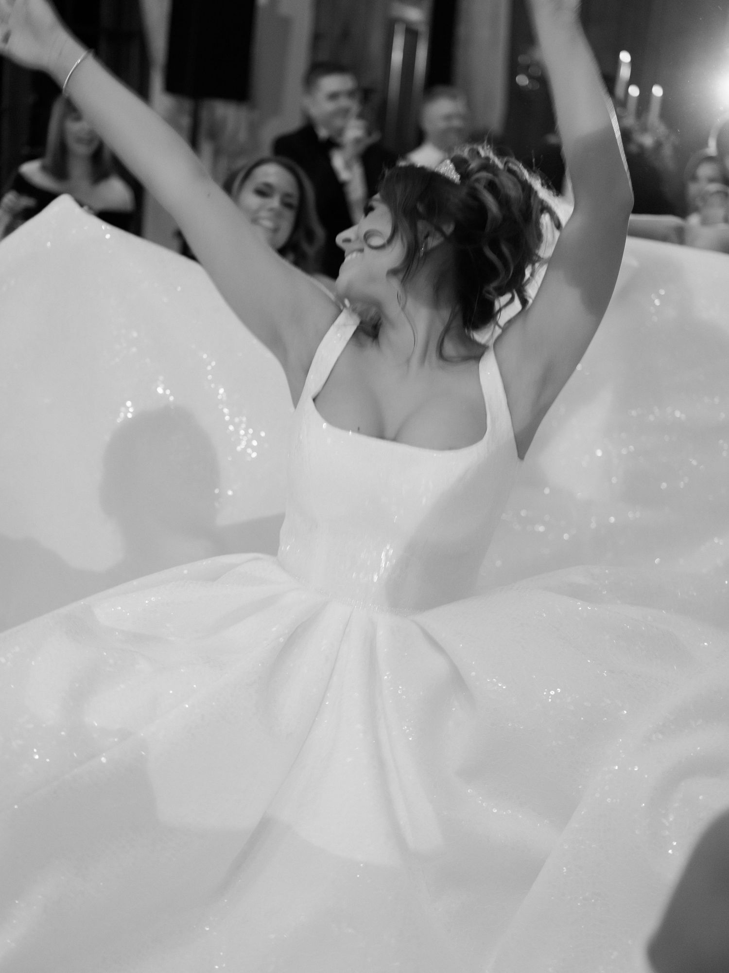 A bride in a sparkling white ballgown twirls joyfully on the dance floor with arms raised during her wedding reception.