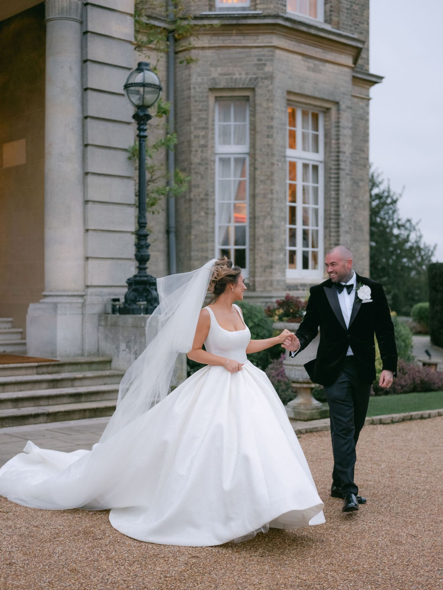 A couple in formal wedding attire walks together on a gravel path outside an elegant stone mansion with vintage lampposts.