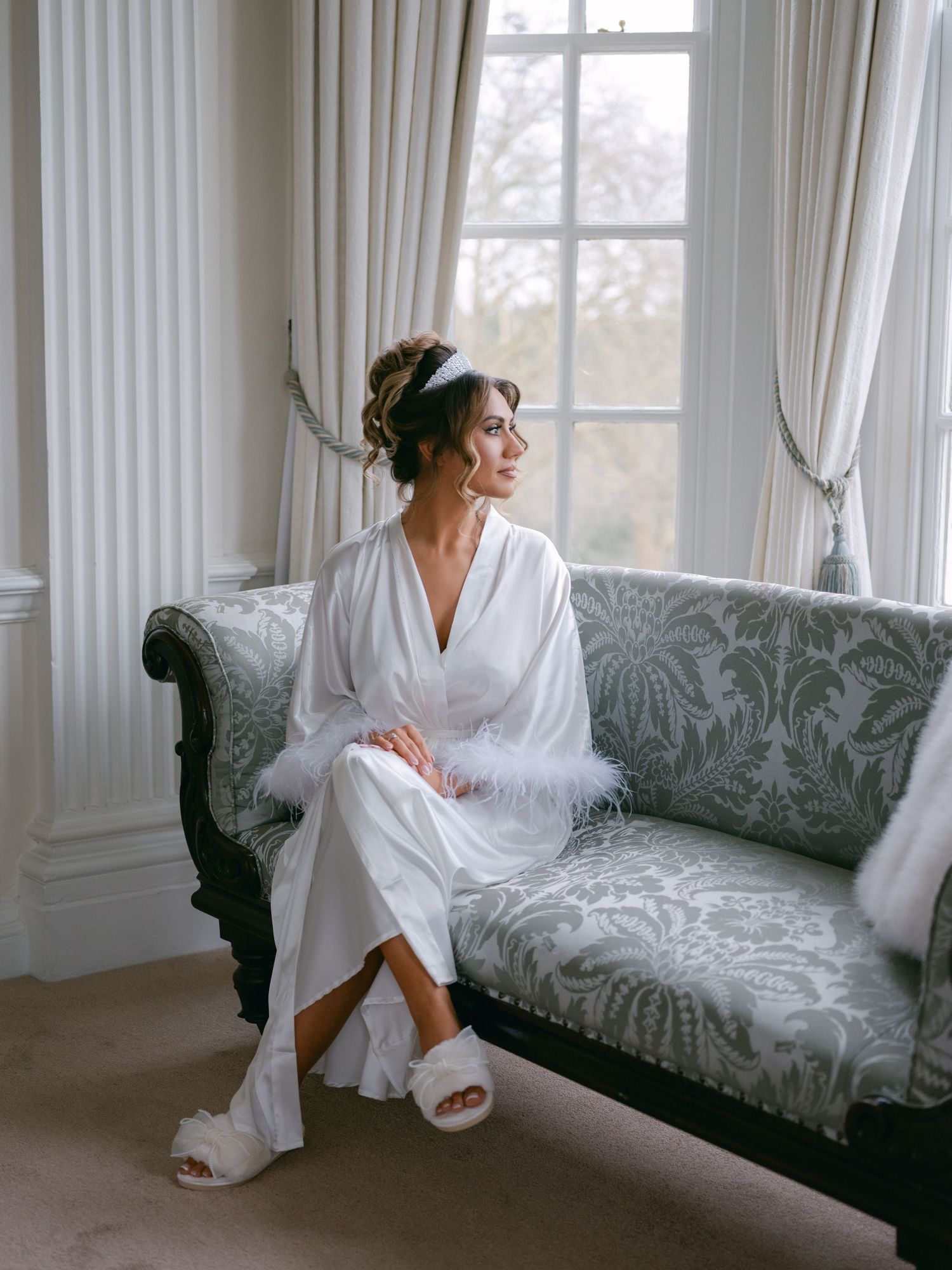 A bride in a white robe and slippers sits elegantly on a gray damask sofa near a bright window with flowing curtains.