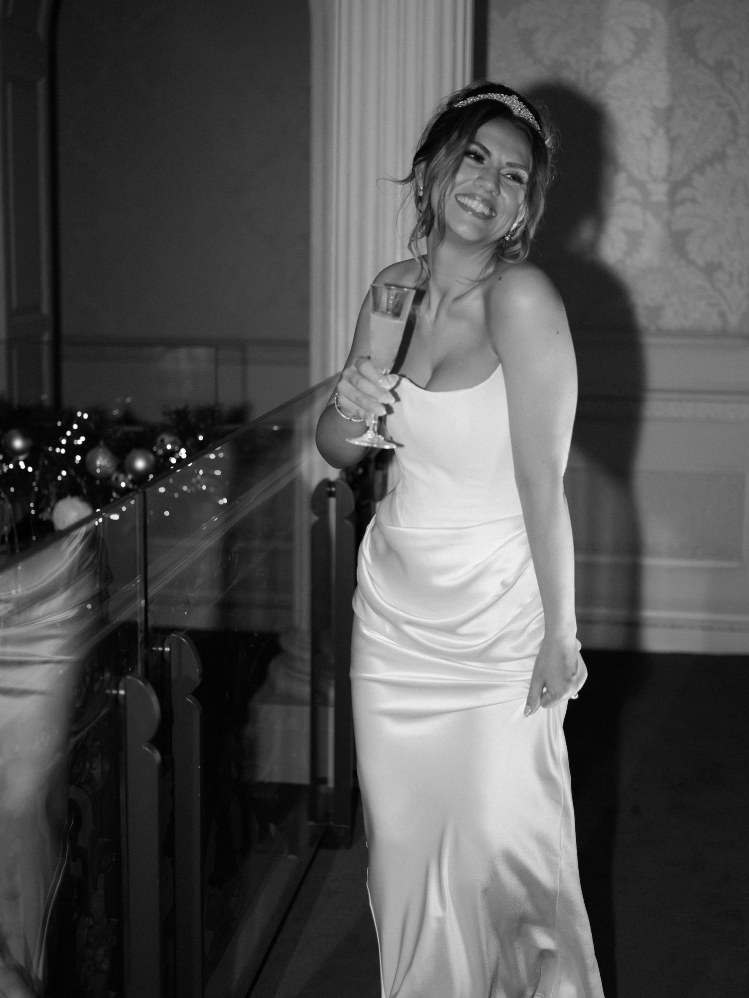 A woman in a flowing white dress poses gracefully in a dimly lit room with twinkling lights in the background.