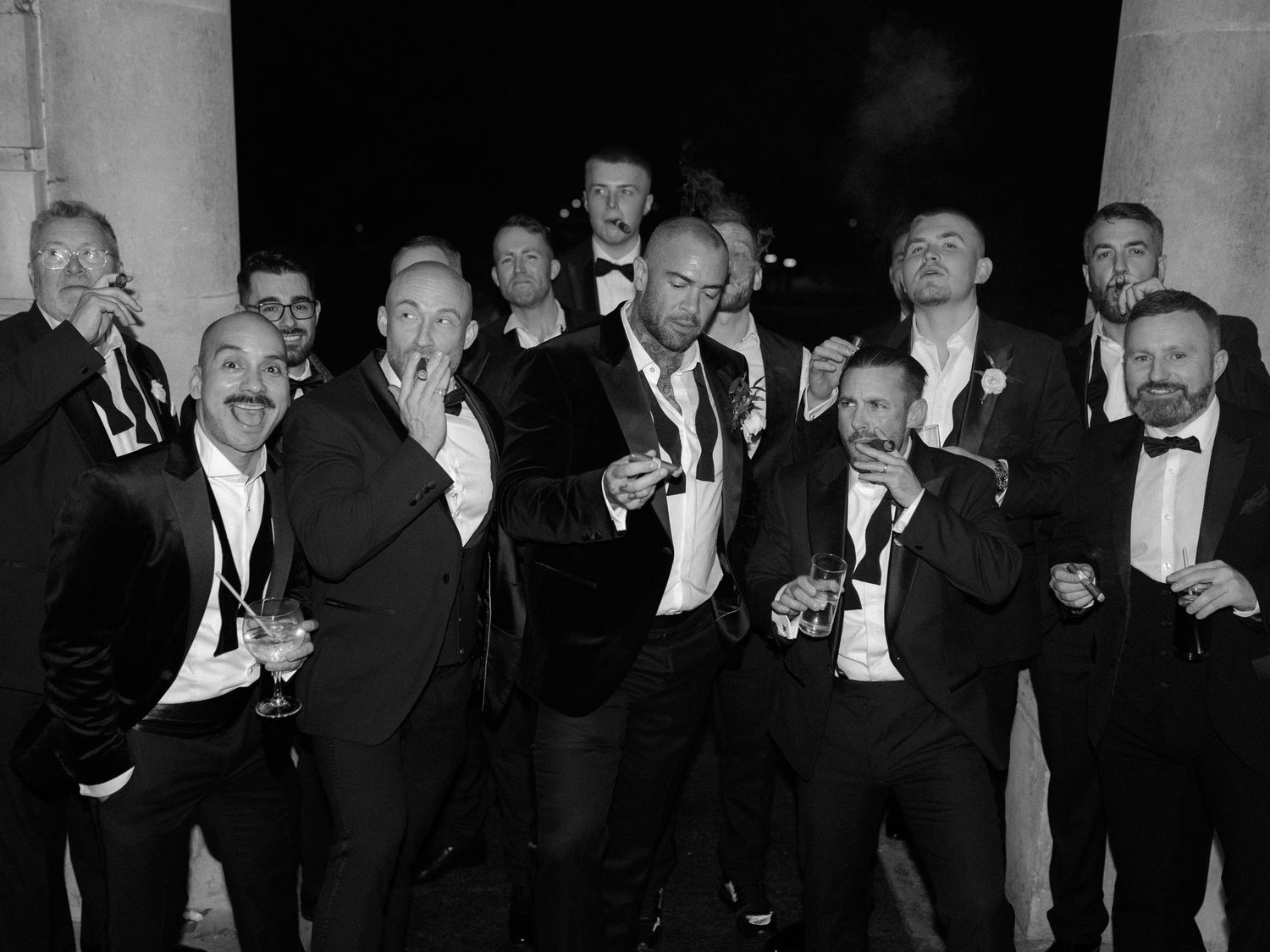 A vintage black and white photo of men in formal attire and bow ties enjoying drinks and cigars at an evening gathering.