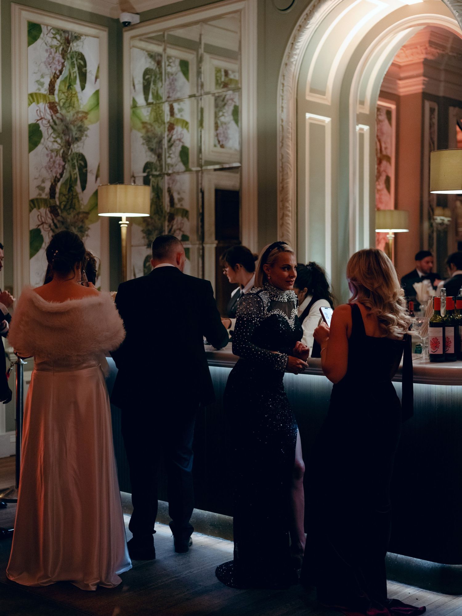 Elegantly dressed dinner guests socialize in a luxurious restaurant with arched doorways and ambient lighting.