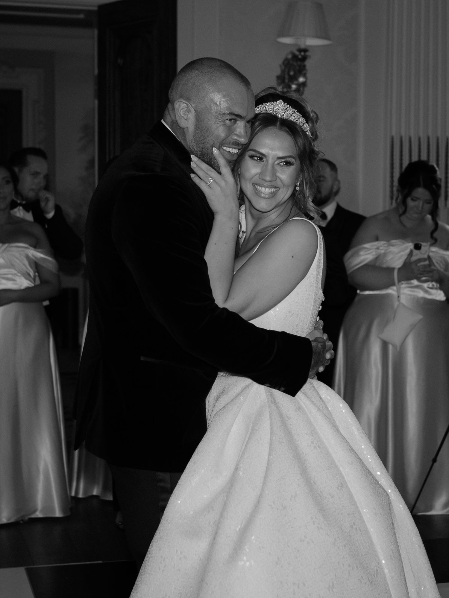 A black and white photo of a couple sharing their first dance at a wedding reception, surrounded by bridesmaids in light dresses.