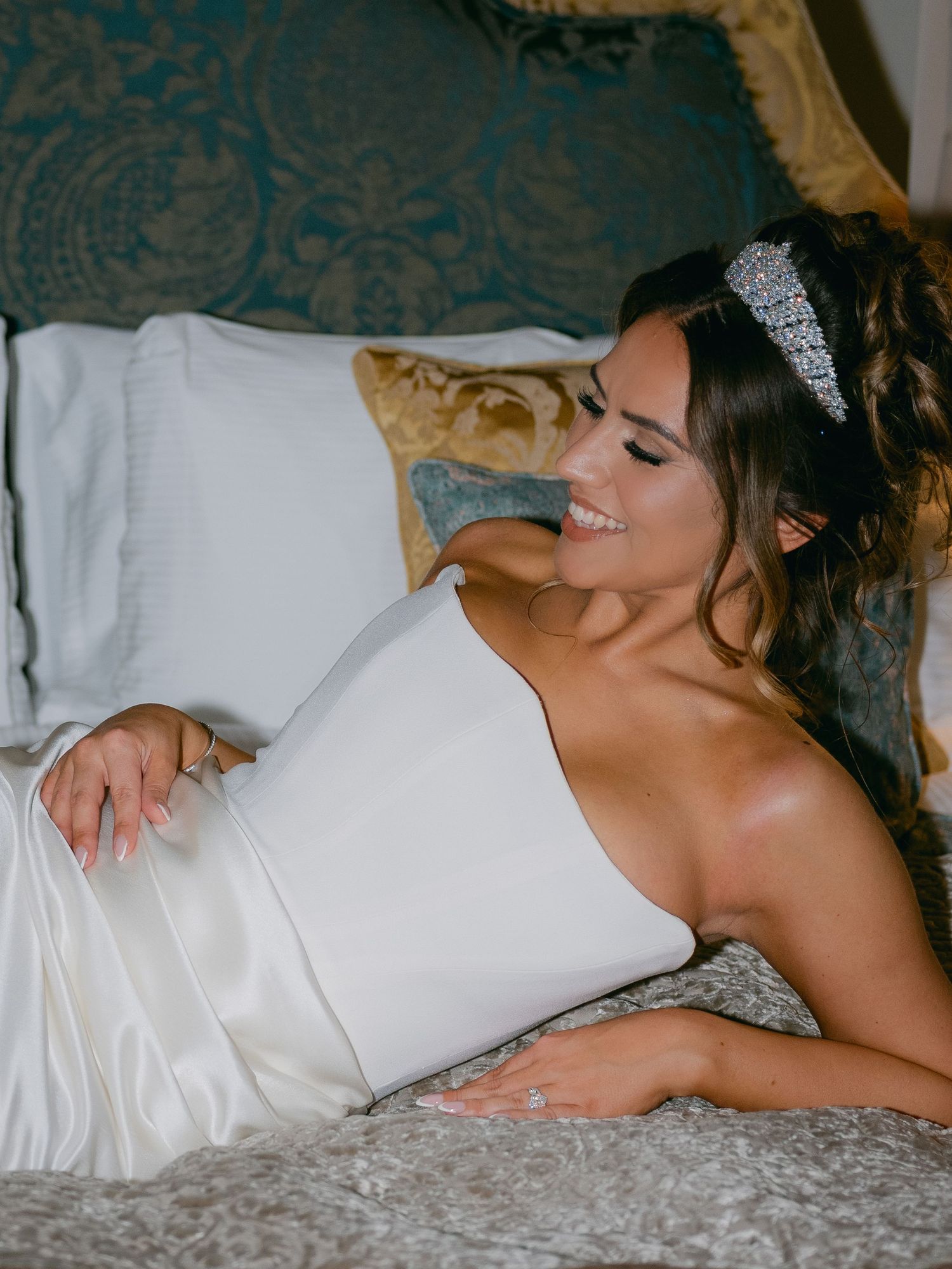 Elegant bride in white one-shoulder gown lounges on luxurious bedding with crystal headpiece and soft romantic lighting.