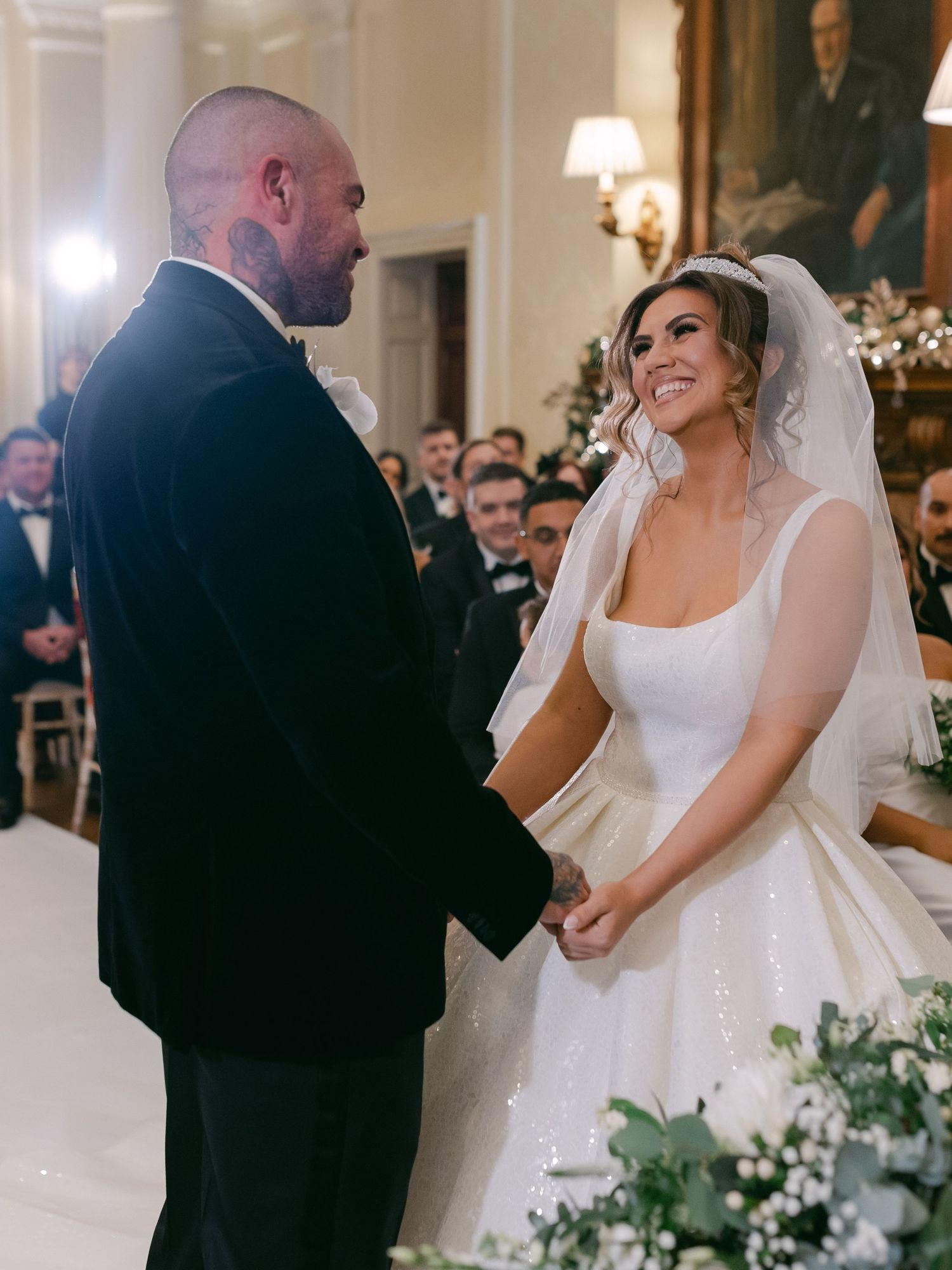 A bride in a white wedding dress and groom in a black suit hold hands during their indoor ceremony ceremony.