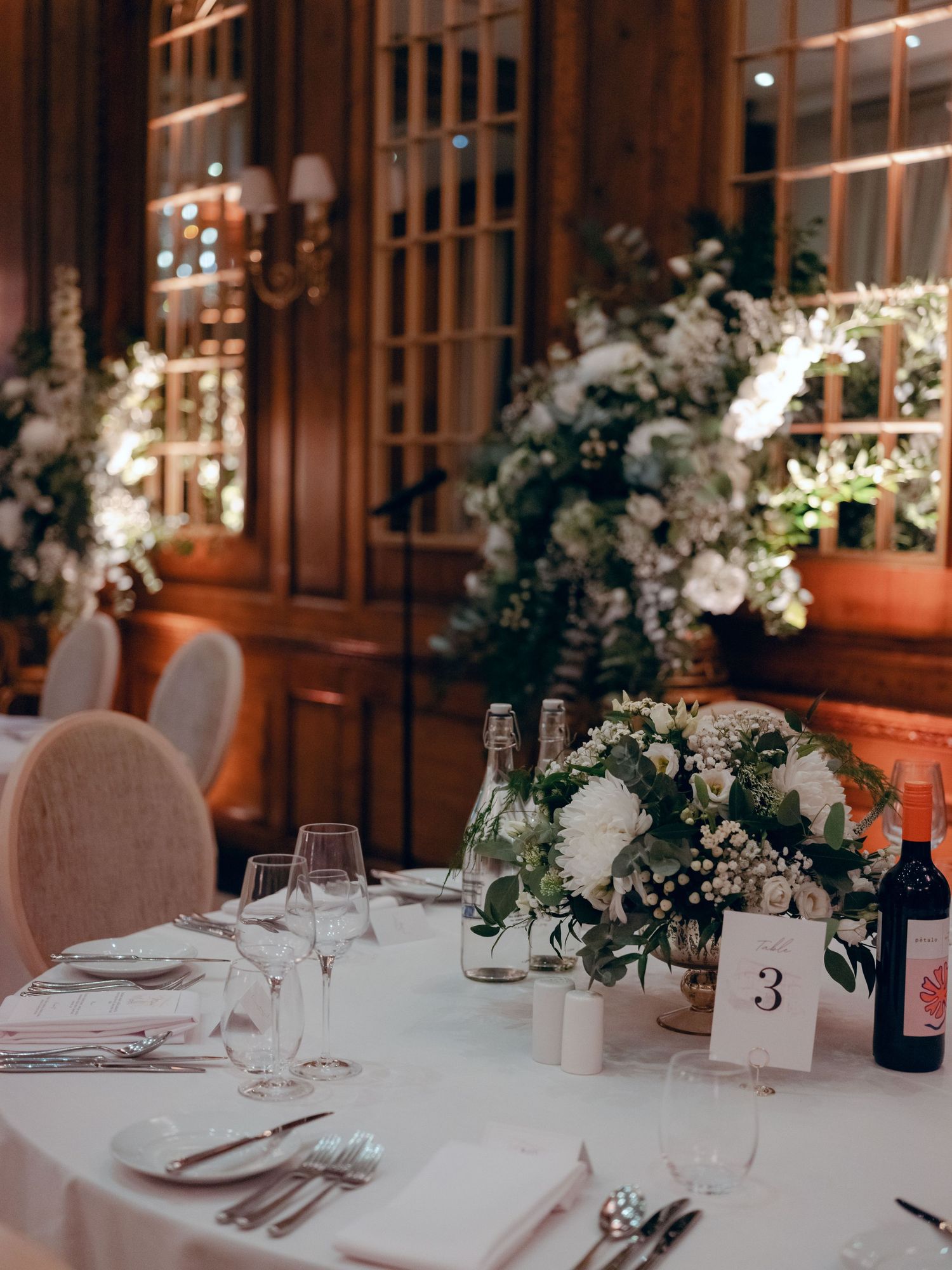 Elegant wedding reception table setting with white floral centerpieces, wine glasses, and table number in a warm wooden room.