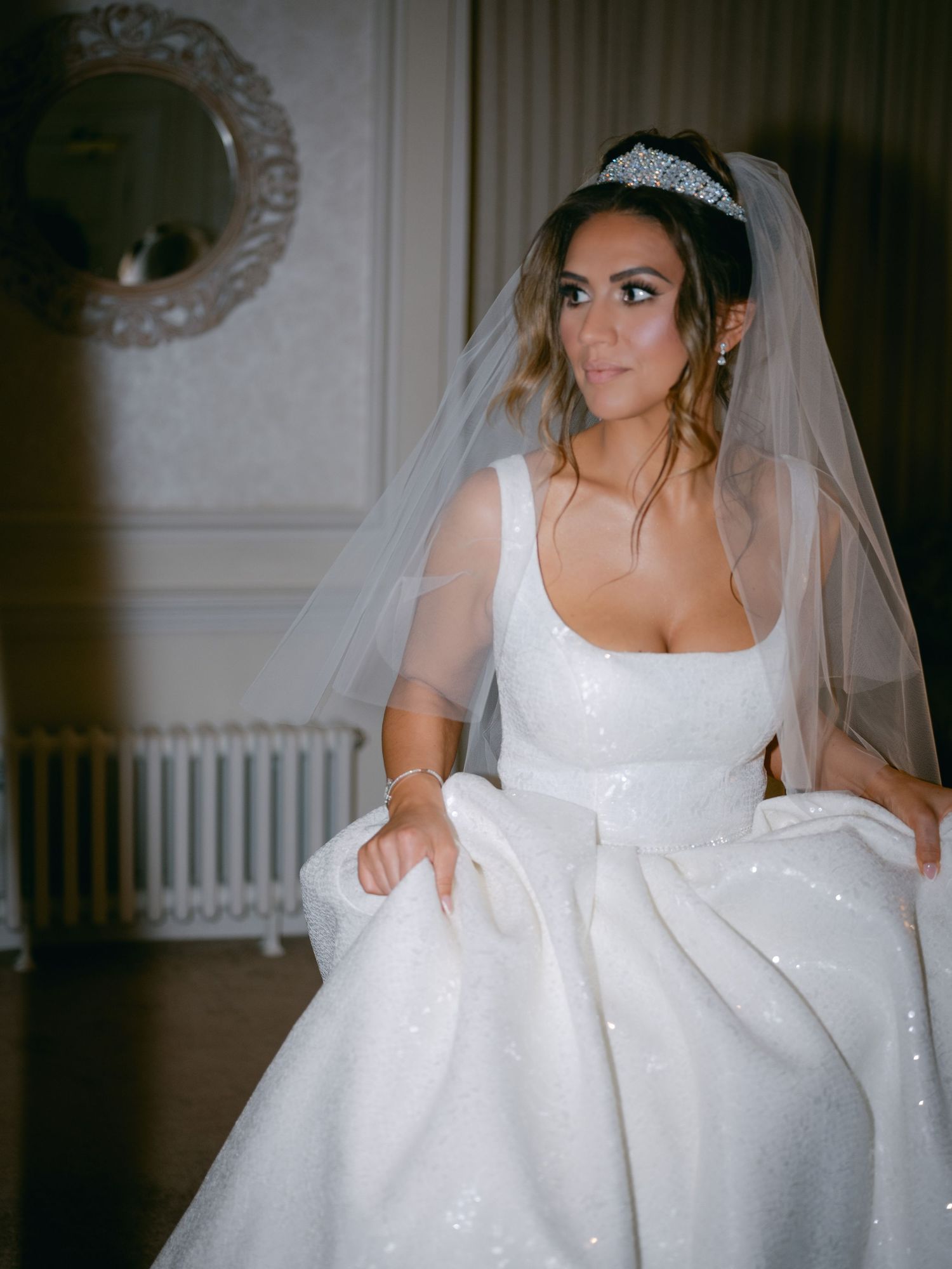 A bride in a sparkly white ballgown wedding dress and veil poses gracefully in an elegant room with ornate details.