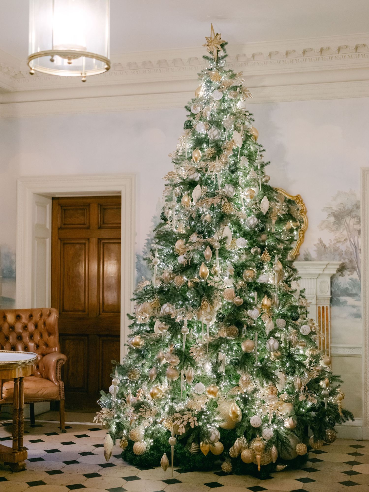 An elegant Christmas tree decorated in white and gold stands in a formal room with wallpaper murals and a patterned floor.