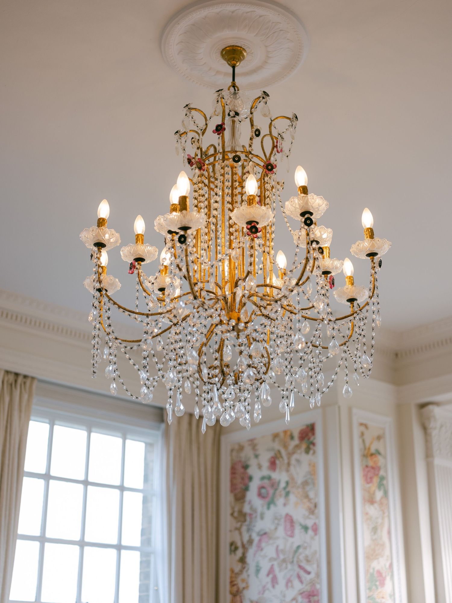 Elegant crystal chandelier with gold metallic frame and multiple tiers of hanging crystals illuminates a room with floral wallpaper.