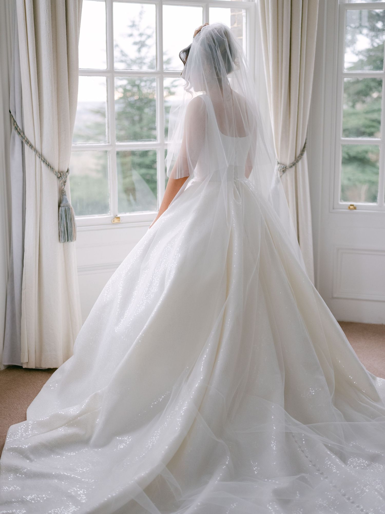 Elegant bride in flowing white ball gown and veil stands before bright window with sheer curtains in natural light.