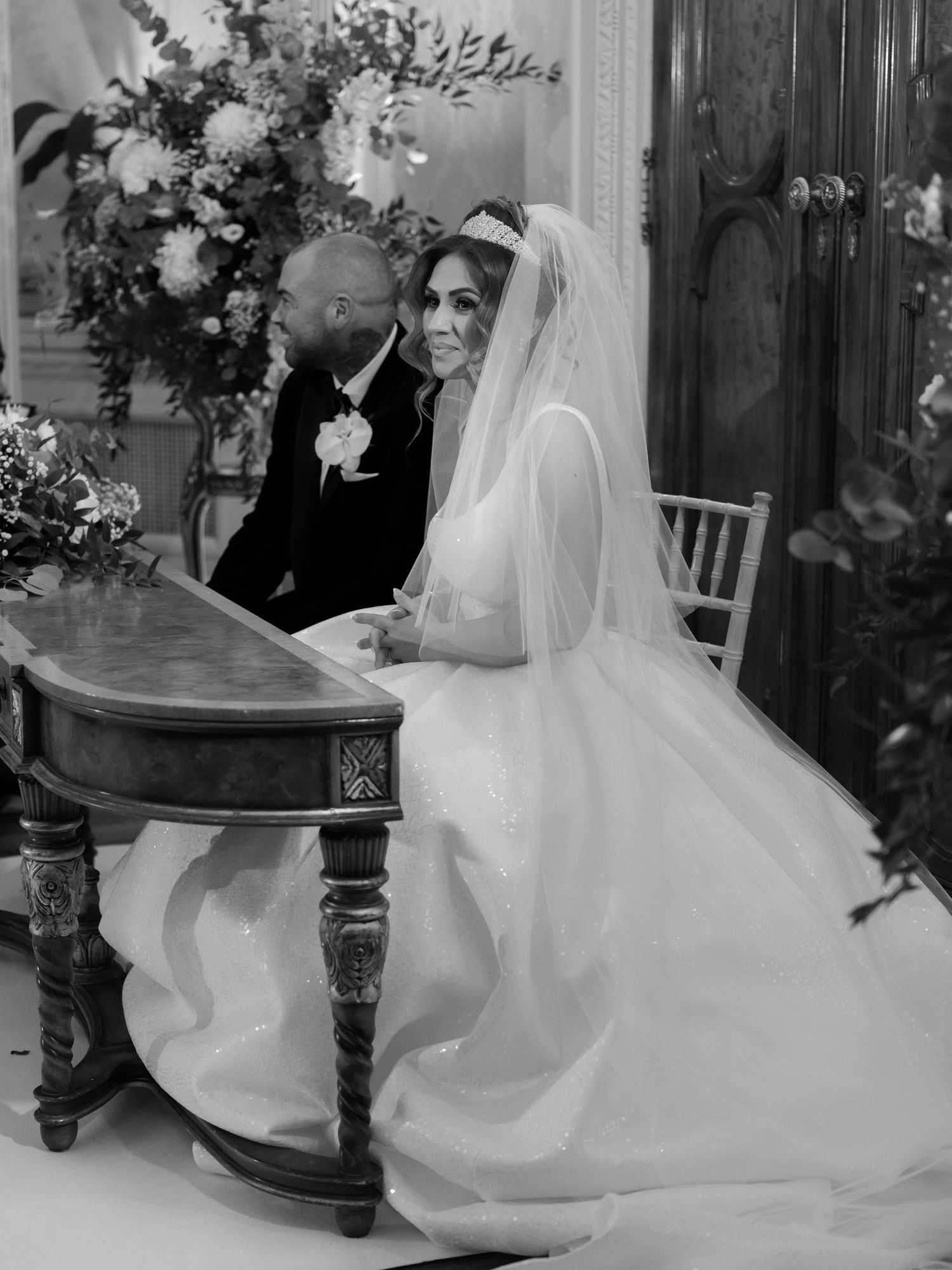 A romantic black and white wedding photograph with a couple sitting at an ornate table surrounded by floral arrangements.
