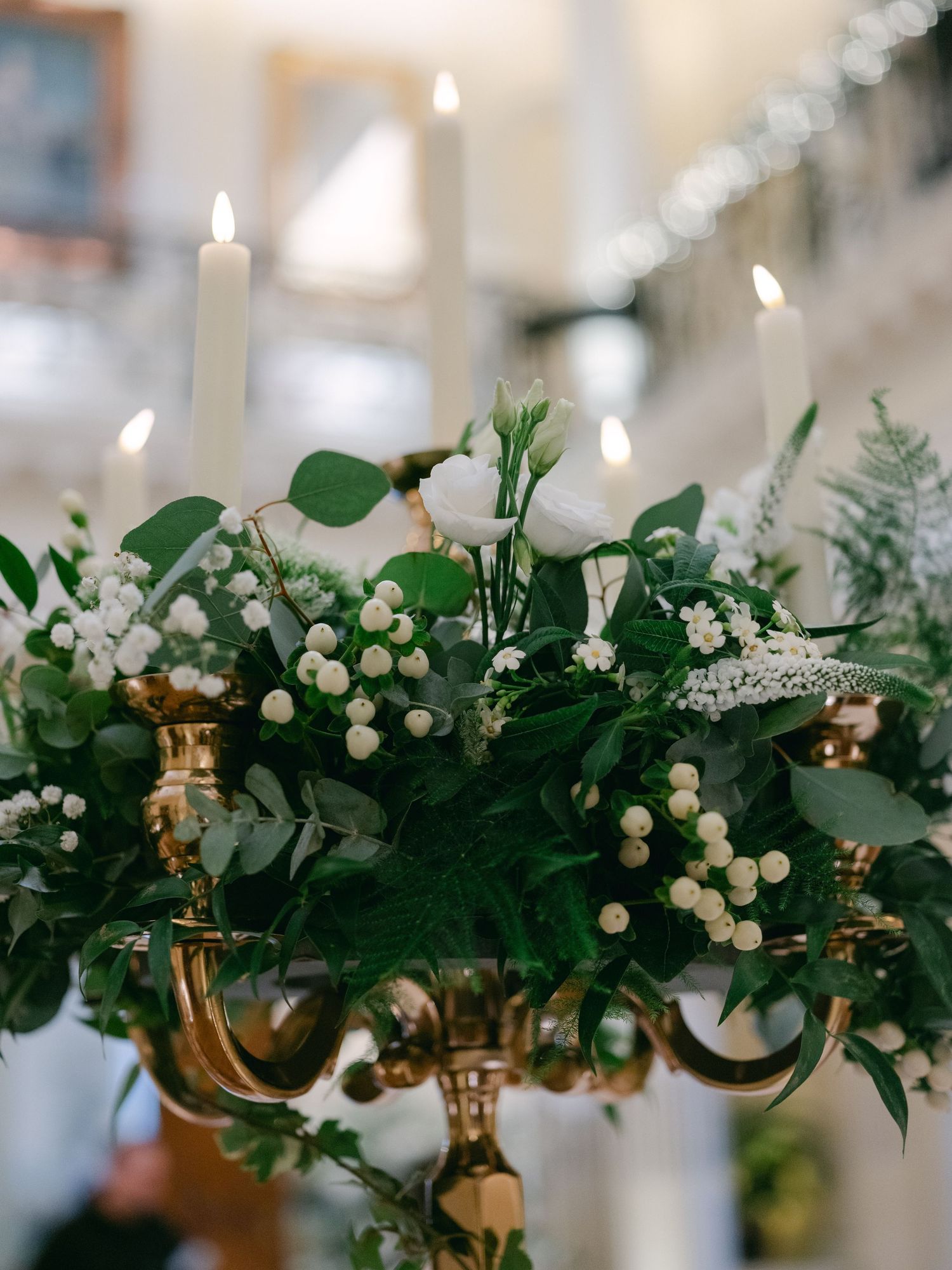 Elegant holiday centerpiece with white candles, berries and greenery arranged on a gold candlestick holder with soft bokeh lights.