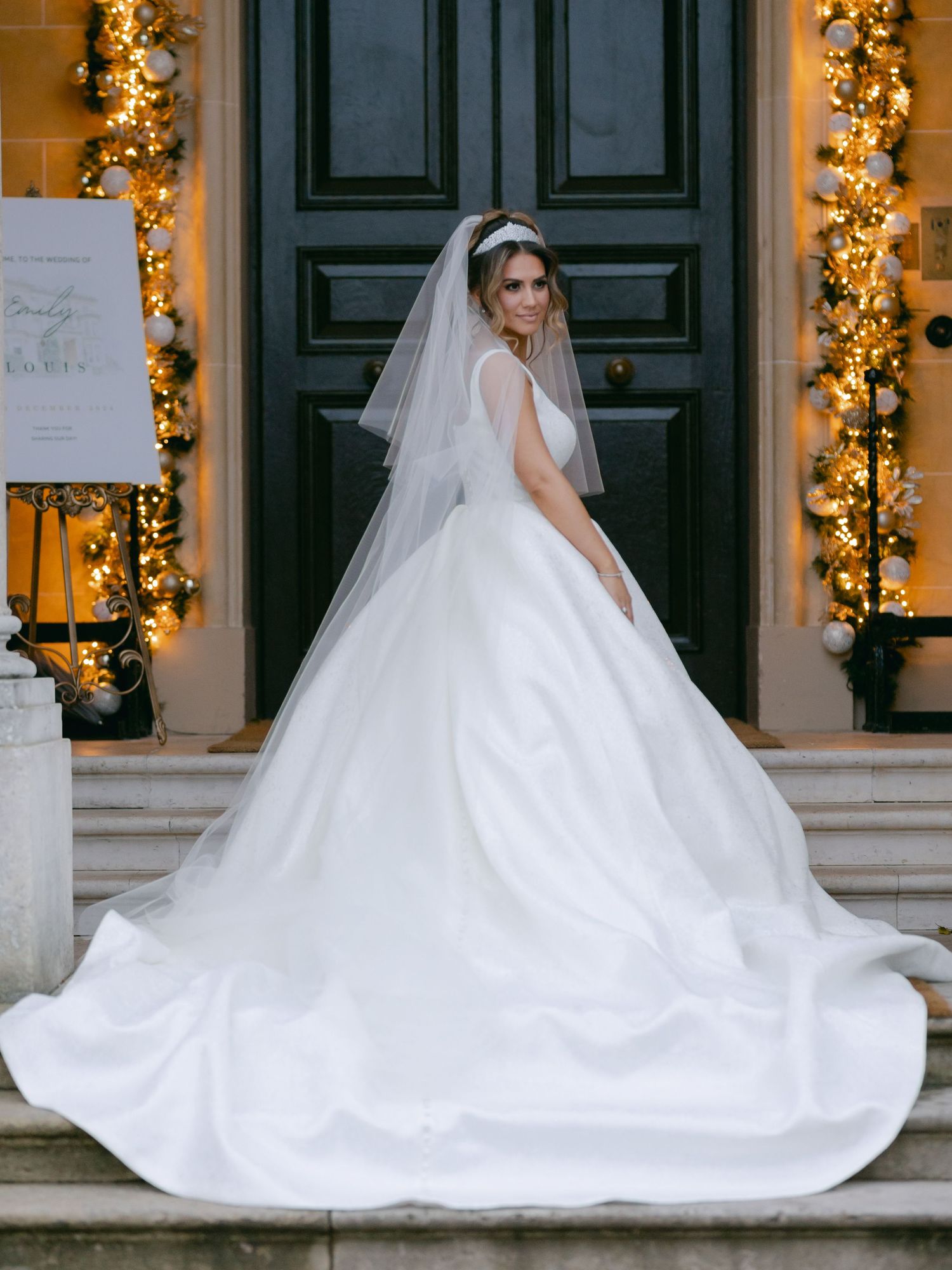 Elegant wedding dress with long train and veil stands on steps decorated with glowing holiday garland and greenery.