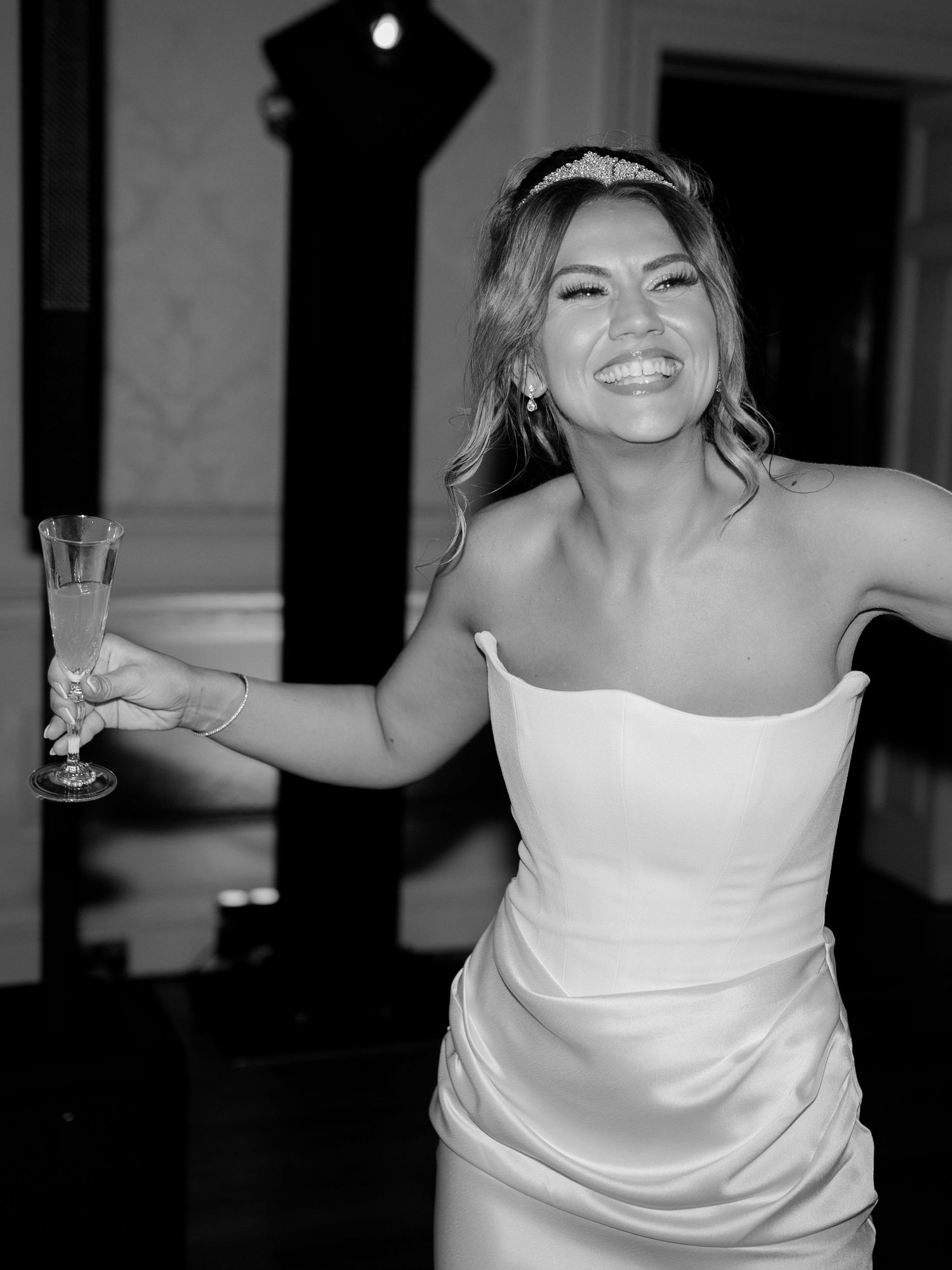 A joyful moment captured in black and white as someone in a strapless white dress raises a champagne glass with a bright smile.