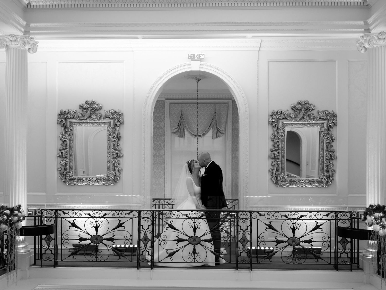 An elegant black and white photograph of a romantic couple embracing on an ornate balcony with decorative mirrors.
