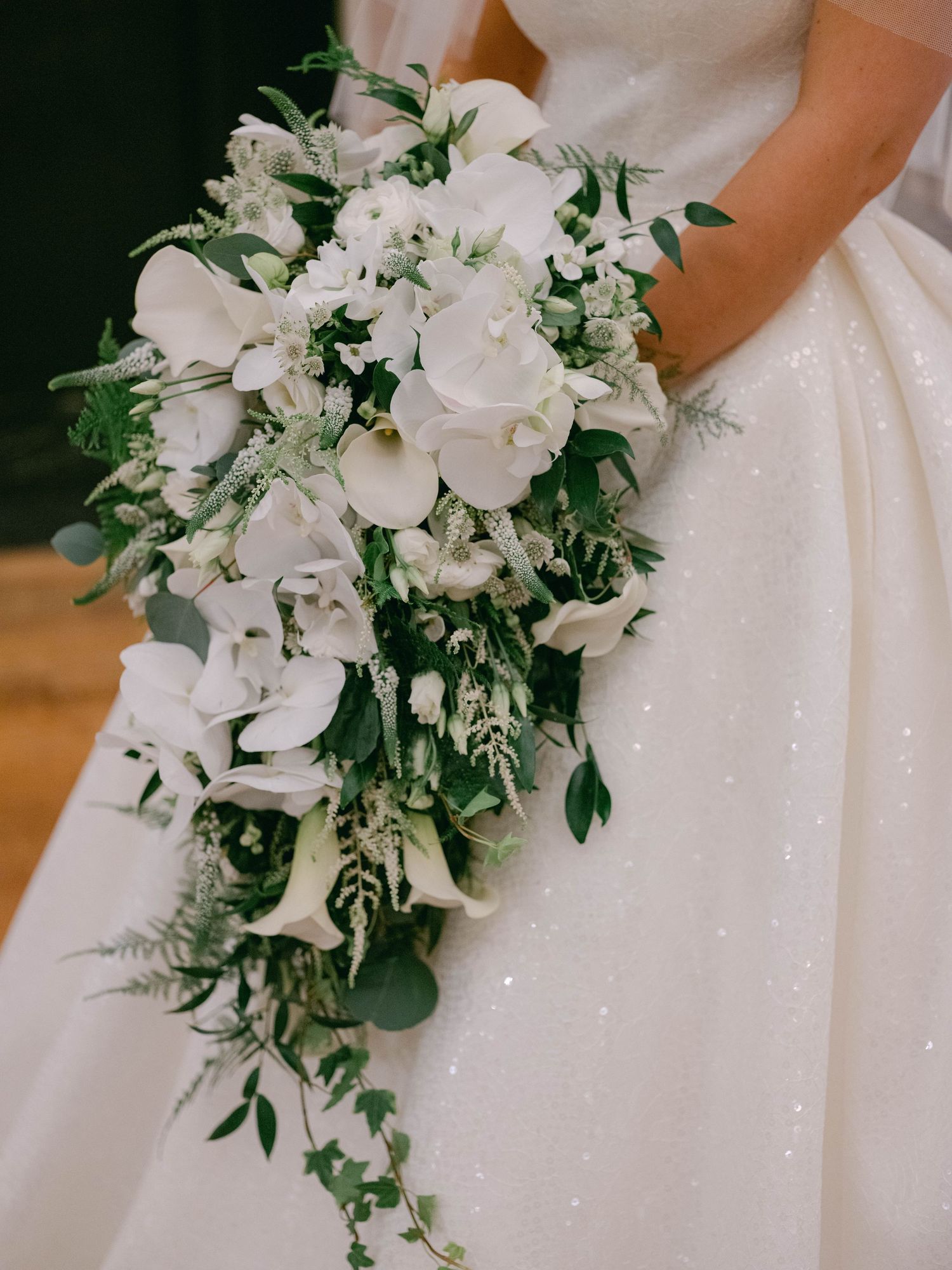 A cascade-style wedding bouquet with white calla lilies, roses, and greenery against a sparkly white dress.